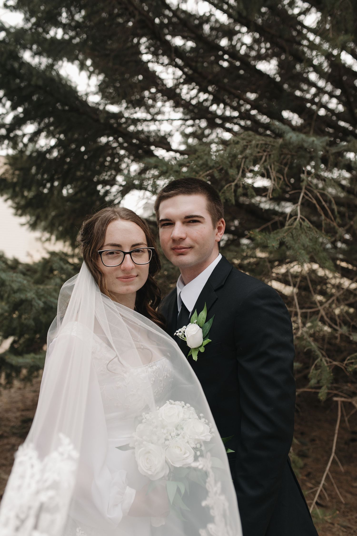 Bride and groom pose together under evergreen trees, bride wearing veil and holding white bouquet.