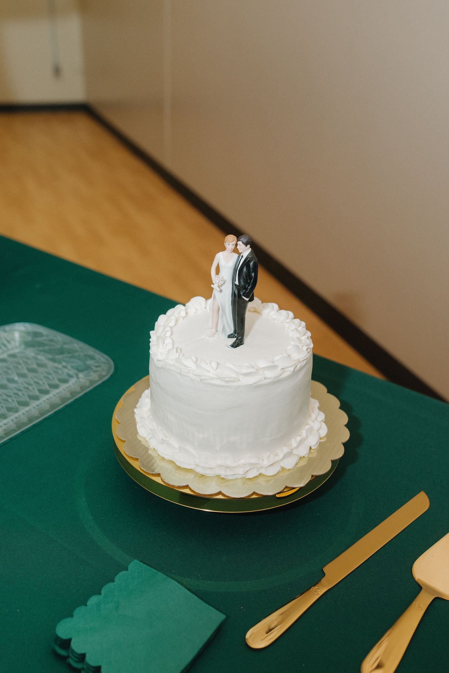 Elegant white wedding cake with bride and groom topper displayed on gold board with green napkins.
