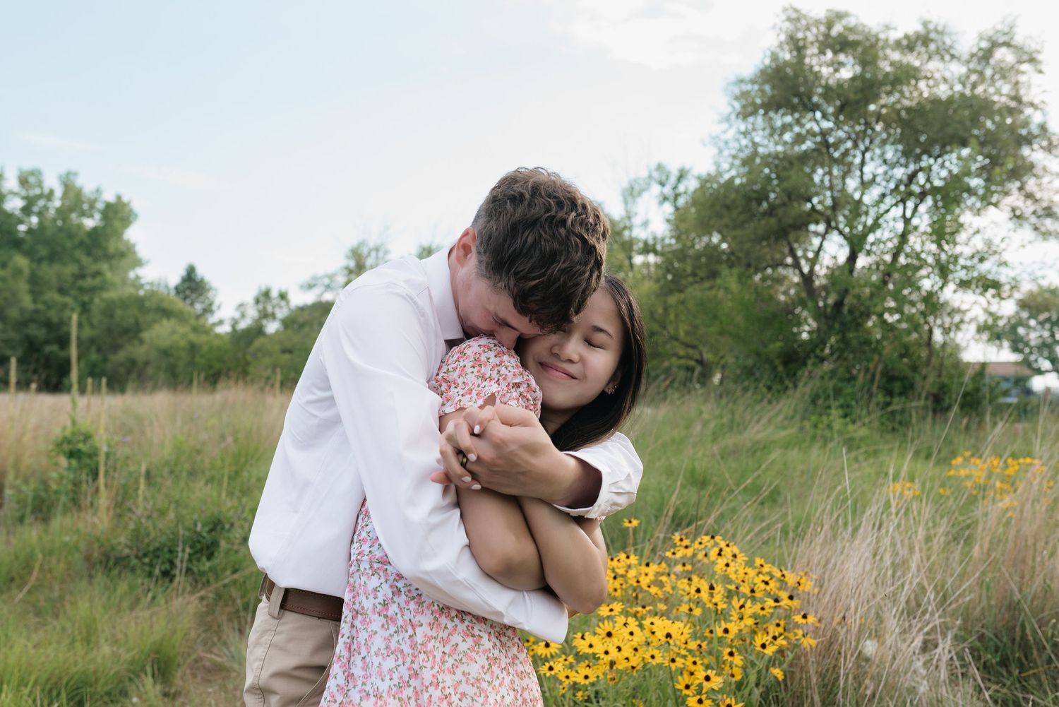 Man hugging woman from behind in field with yellow wildflowers, couple sharing tender moment.
