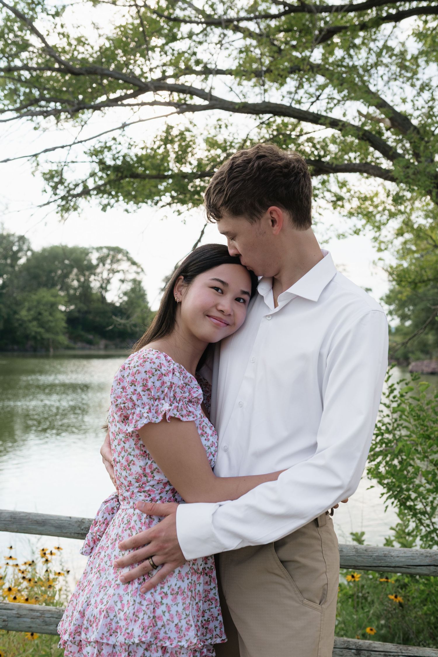 A couple shares a tender embrace near a serene lake surrounded by lush green trees.