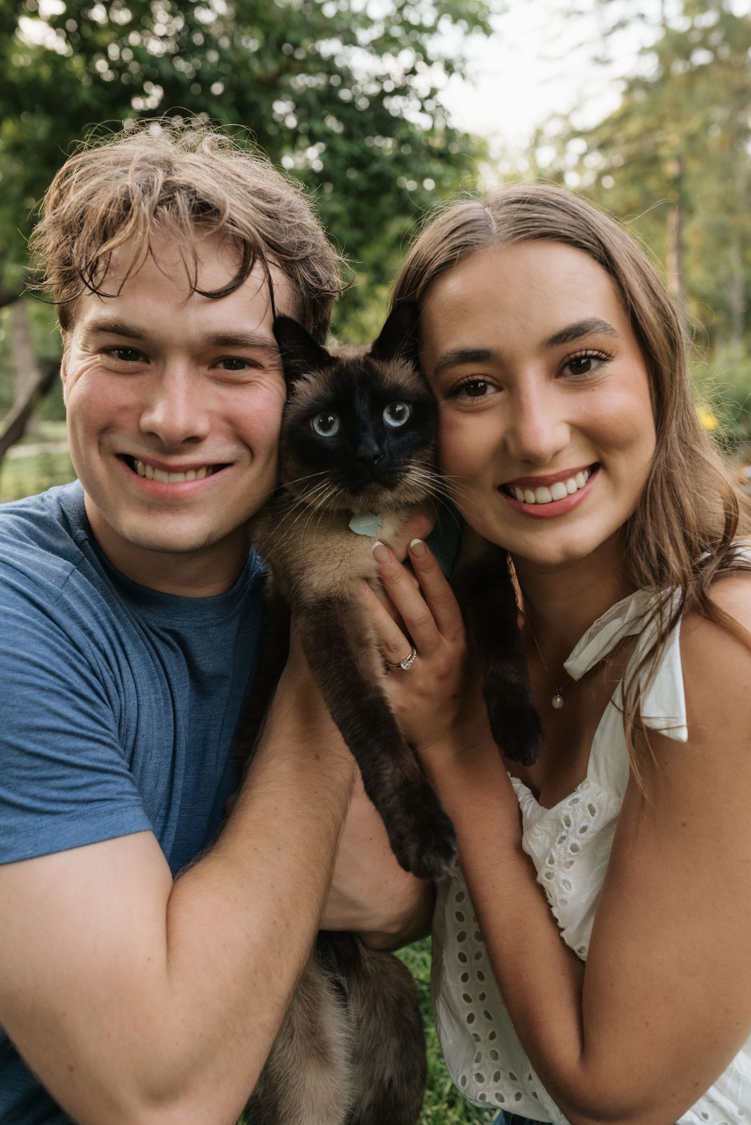 Smiling young couple holding a Siamese cat outdoors in a lush green garden setting.