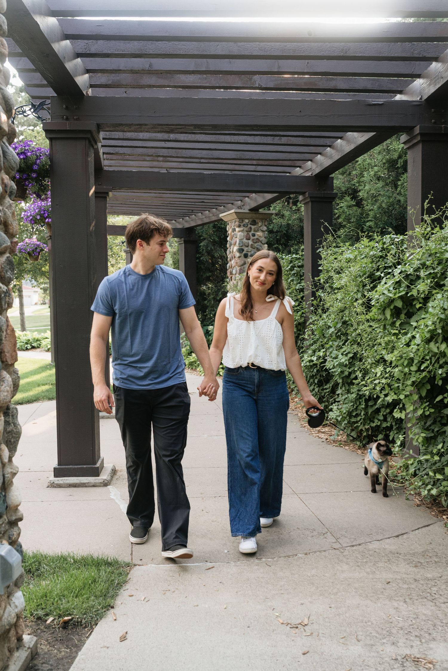 Couple holding hands walking under pergola with dog on leash surrounded by lush greenery.