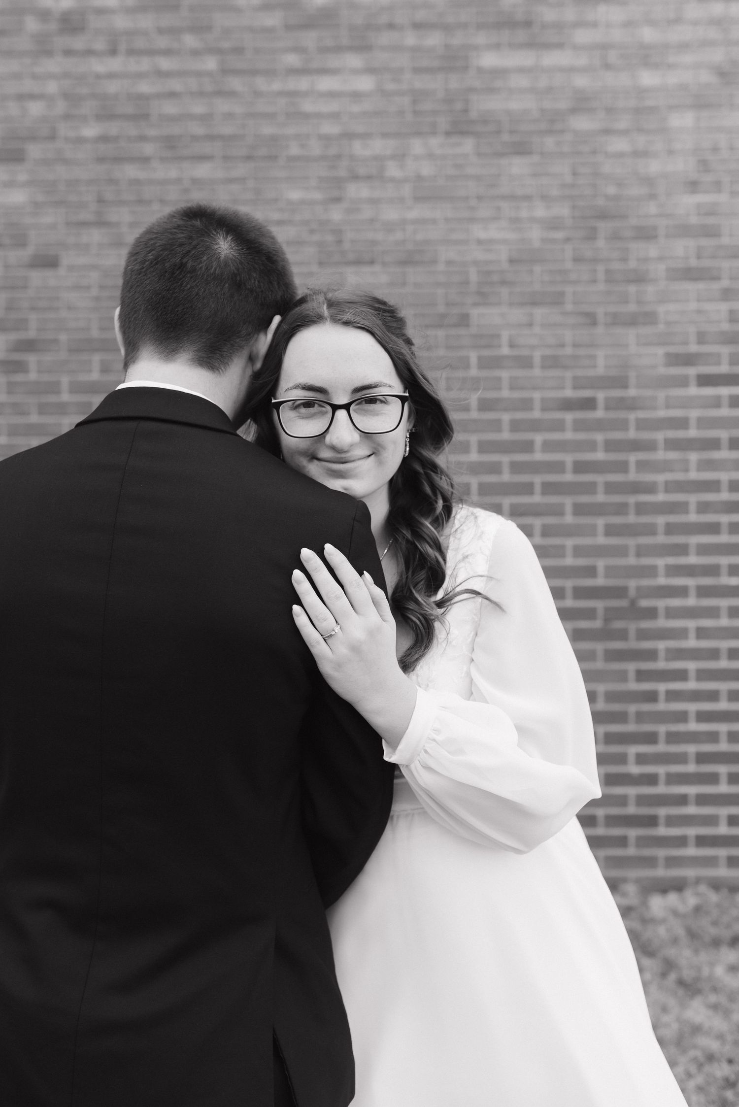 Black and white wedding photo of bride in glasses hugging groom near brick wall, showing ring on hand.
