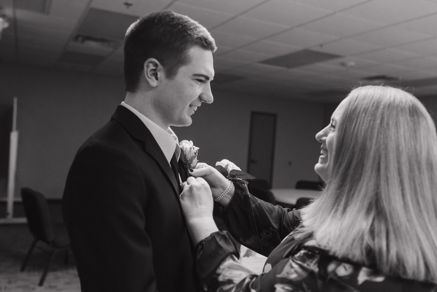 A woman pins a boutonniere on a young man in a black suit at a wedding ceremony.