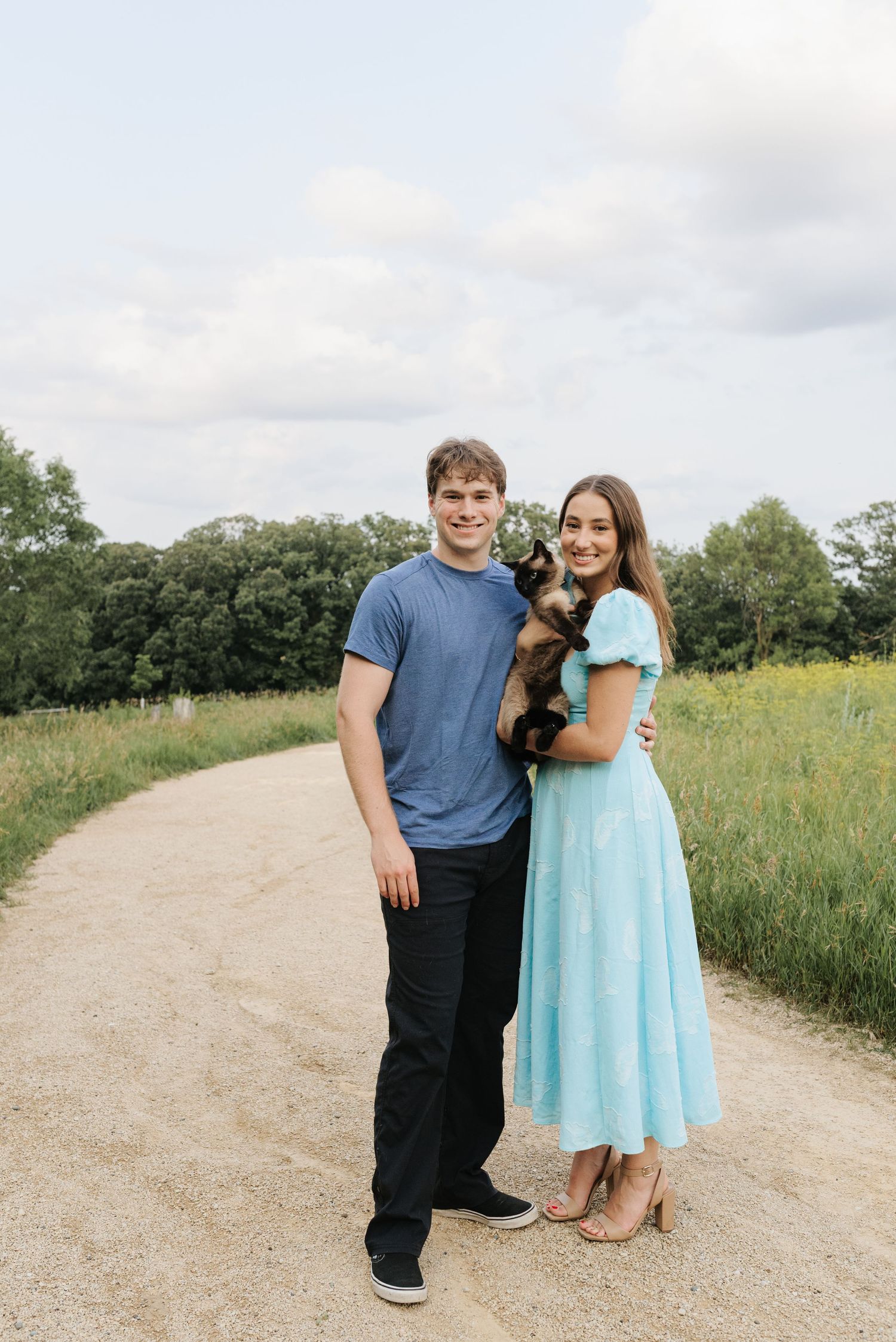 A smiling couple stands on a dirt path holding a small dog, with green trees and a cloudy sky in the background.