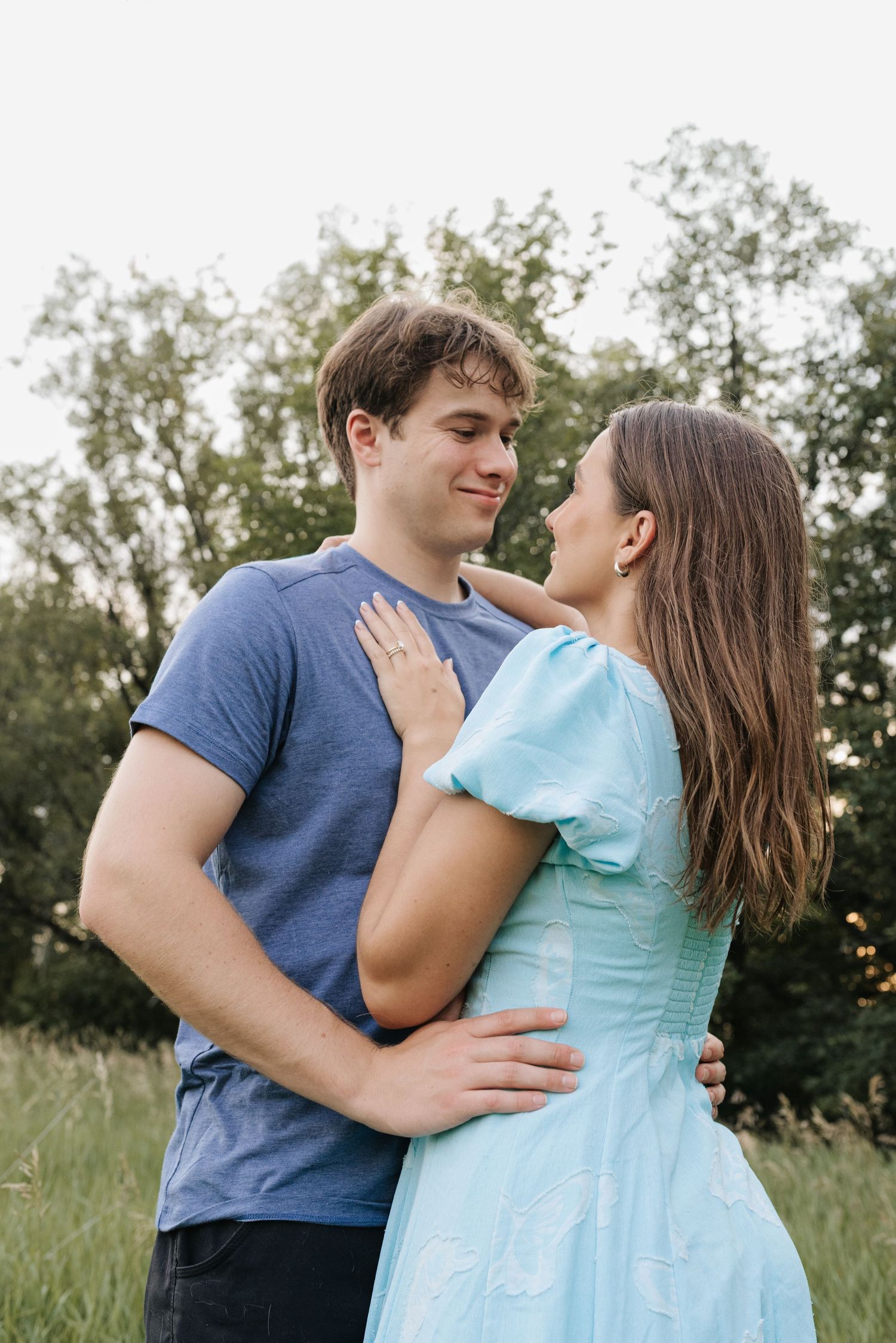 A young couple in a green field, man in blue t-shirt and woman in light blue dress, gazing at each other romantically.