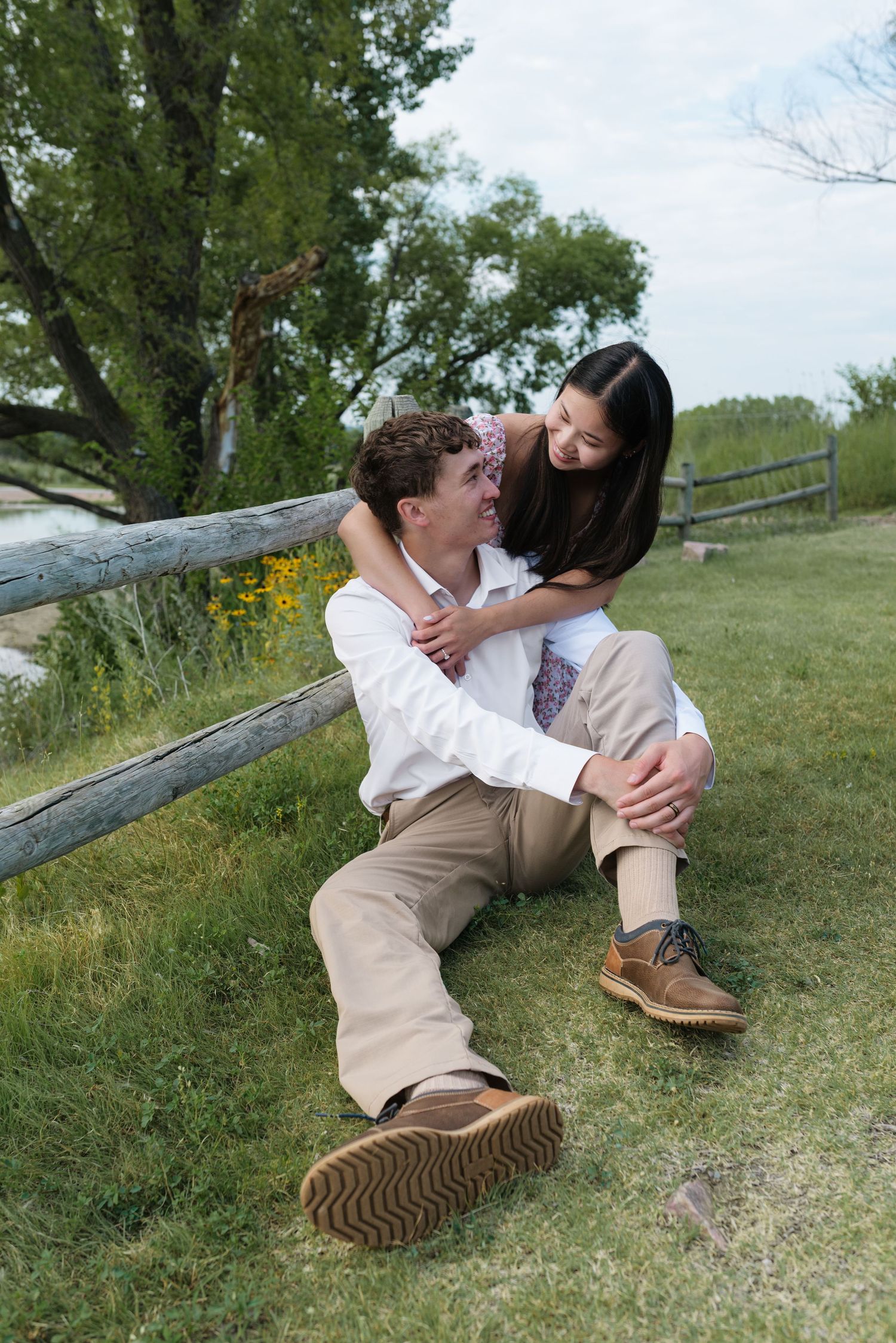 Couple sitting on grass by wooden fence, woman embracing man from behind, smiling outdoors.