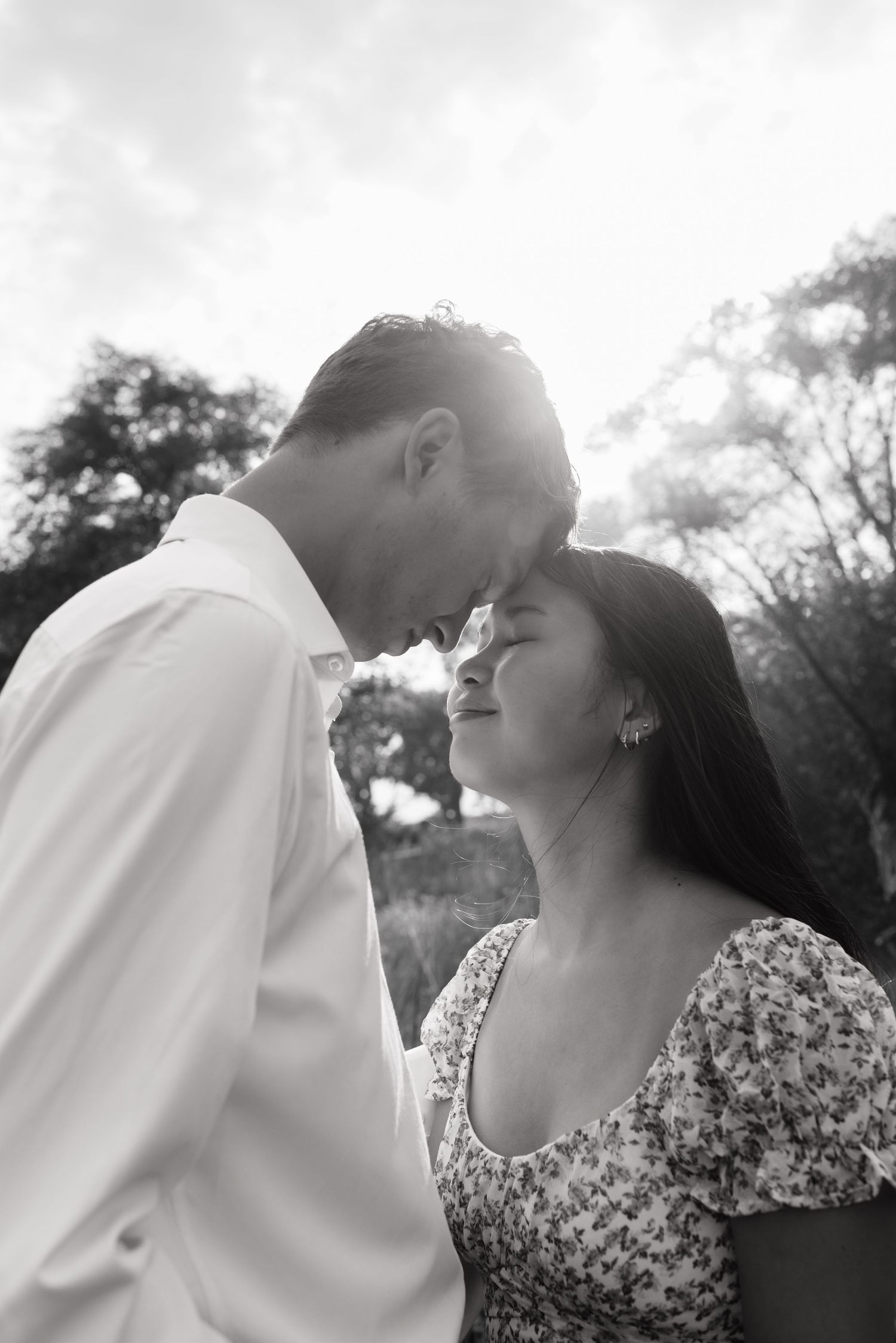 Black and white romantic photo of a couple touching foreheads with eyes closed in soft sunlight.