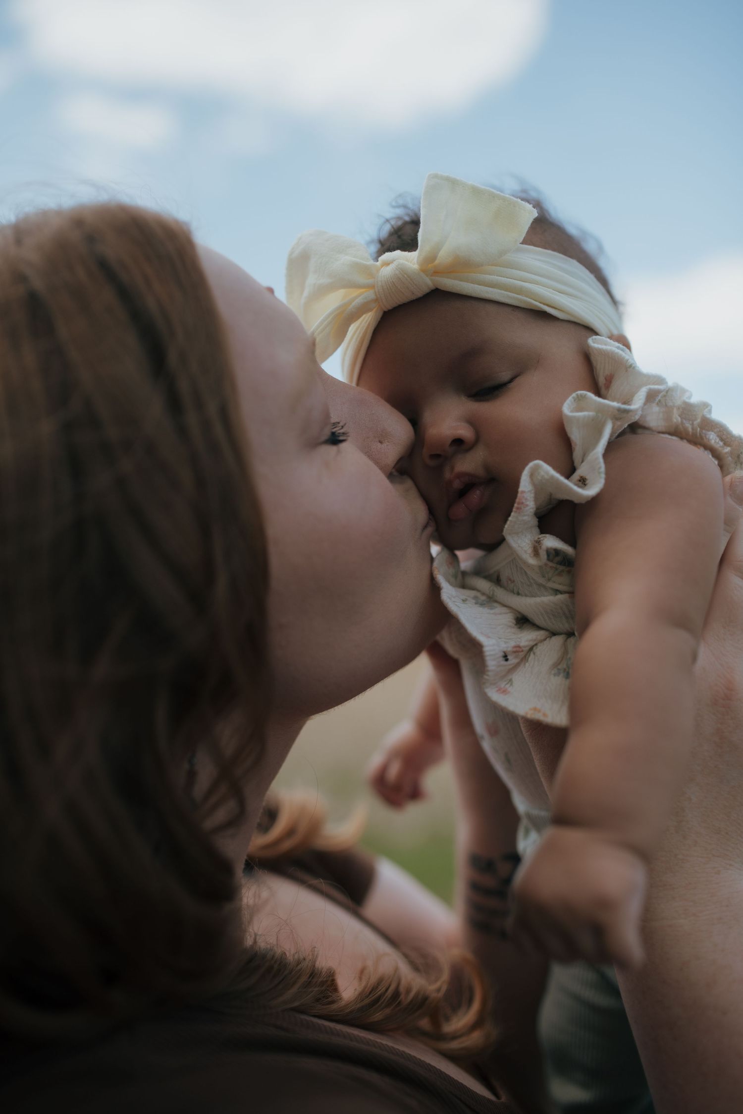 Tender moment of mother kissing newborn baby girl in white bow headband outdoors.