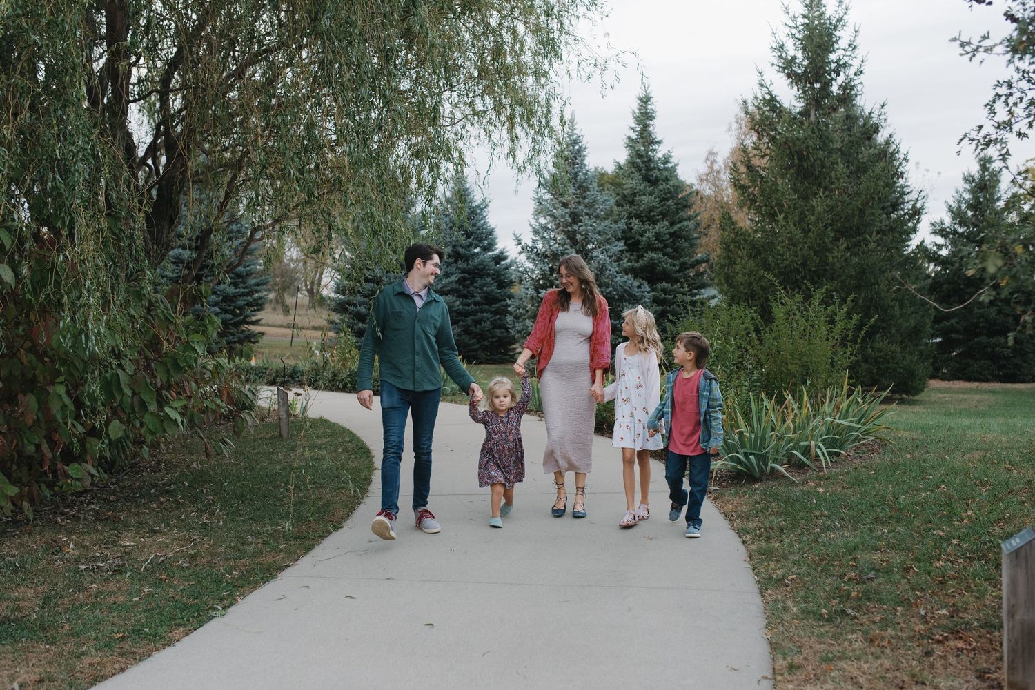 Happy family strolling along a curved park pathway with tall evergreen trees.