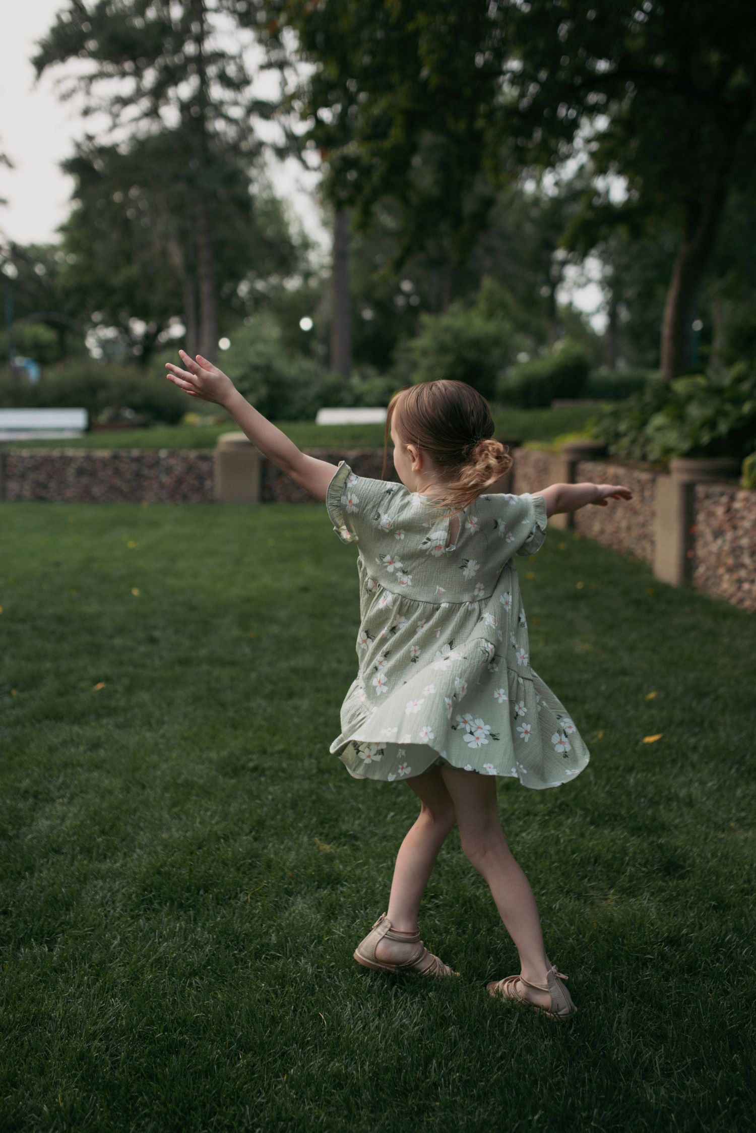 A young girl in a mint floral dress twirls with arms outstretched on green grass in a park.