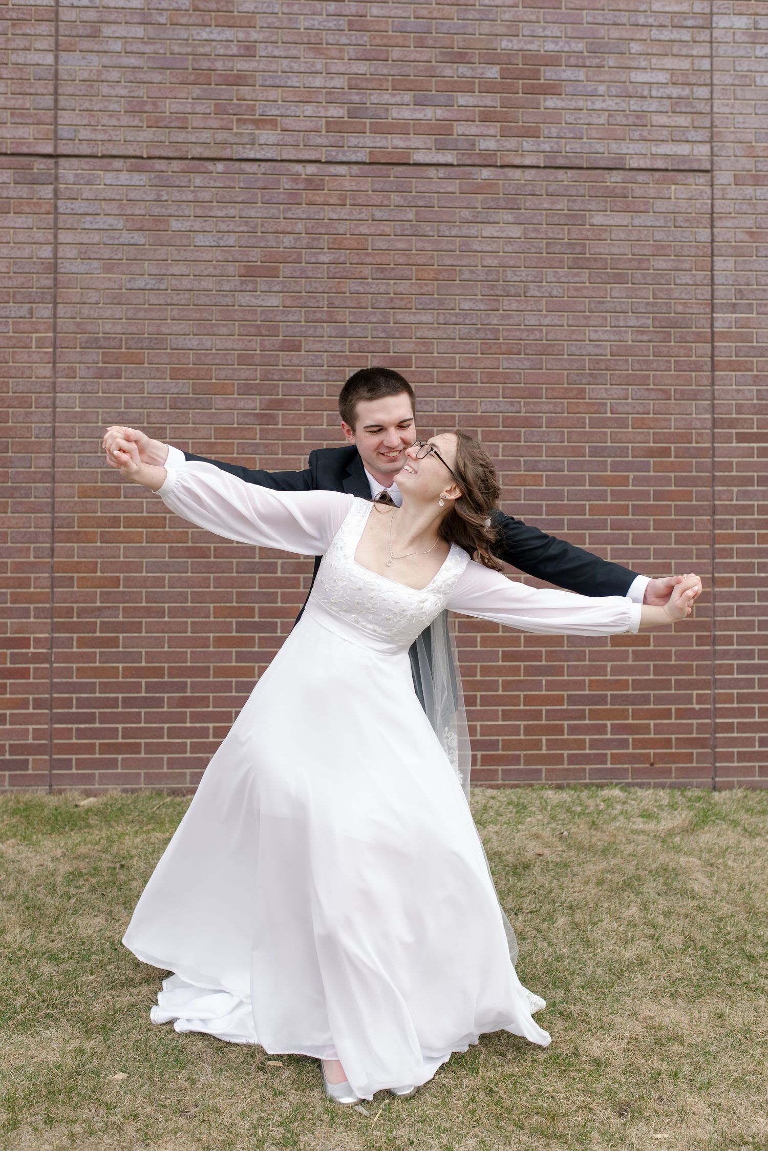 Joyful bride and groom dancing playfully with arms outstretched in front of a red brick wall.