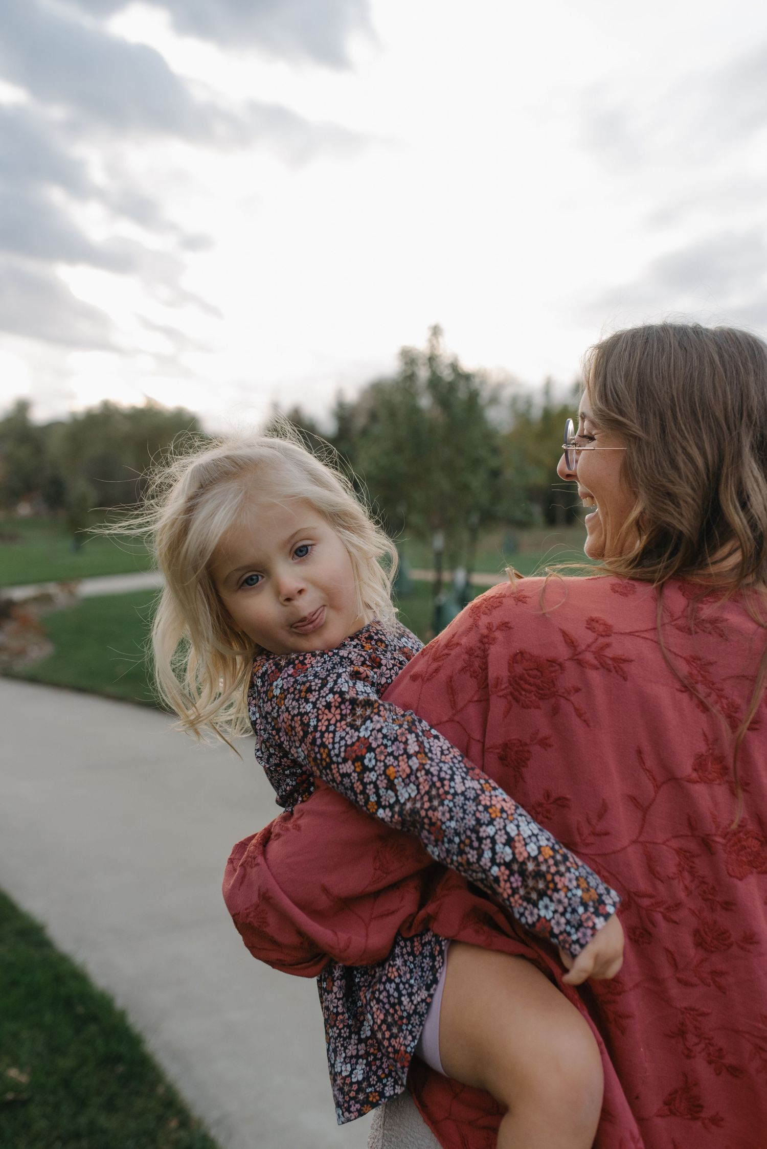A laughing woman carries a playful blonde toddler making a pouty face outdoors.