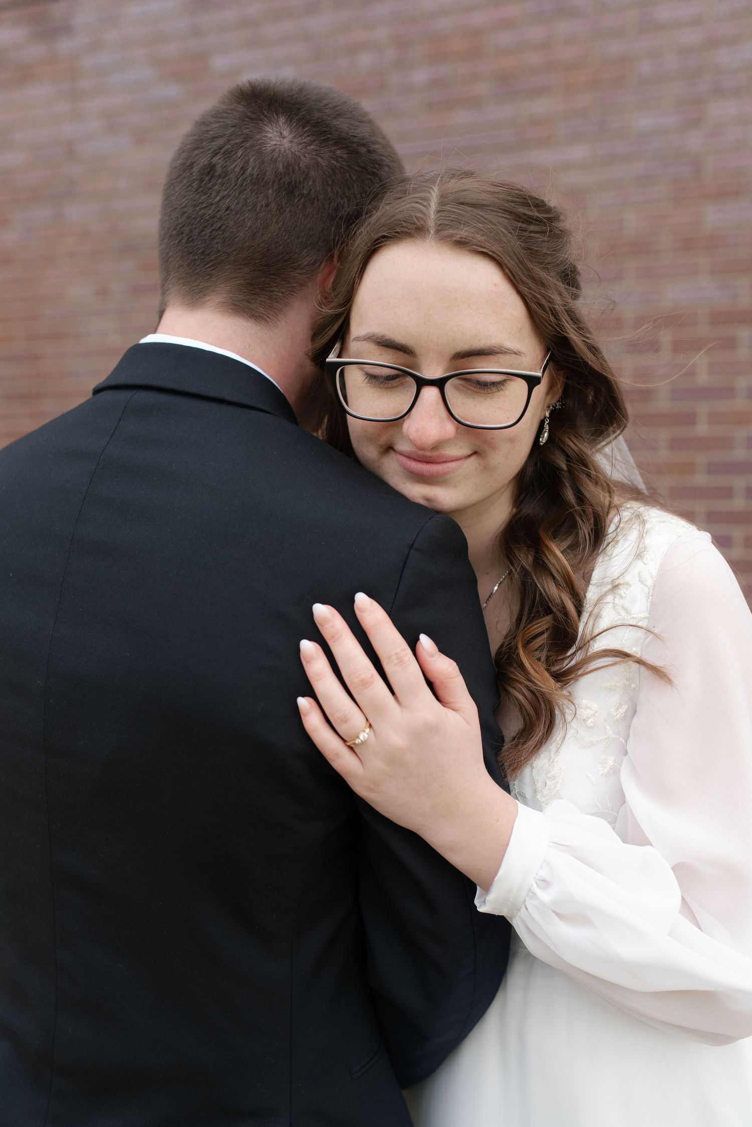 Emotional bride in white lace gown and glasses embraces groom in black suit during wedding ceremony.