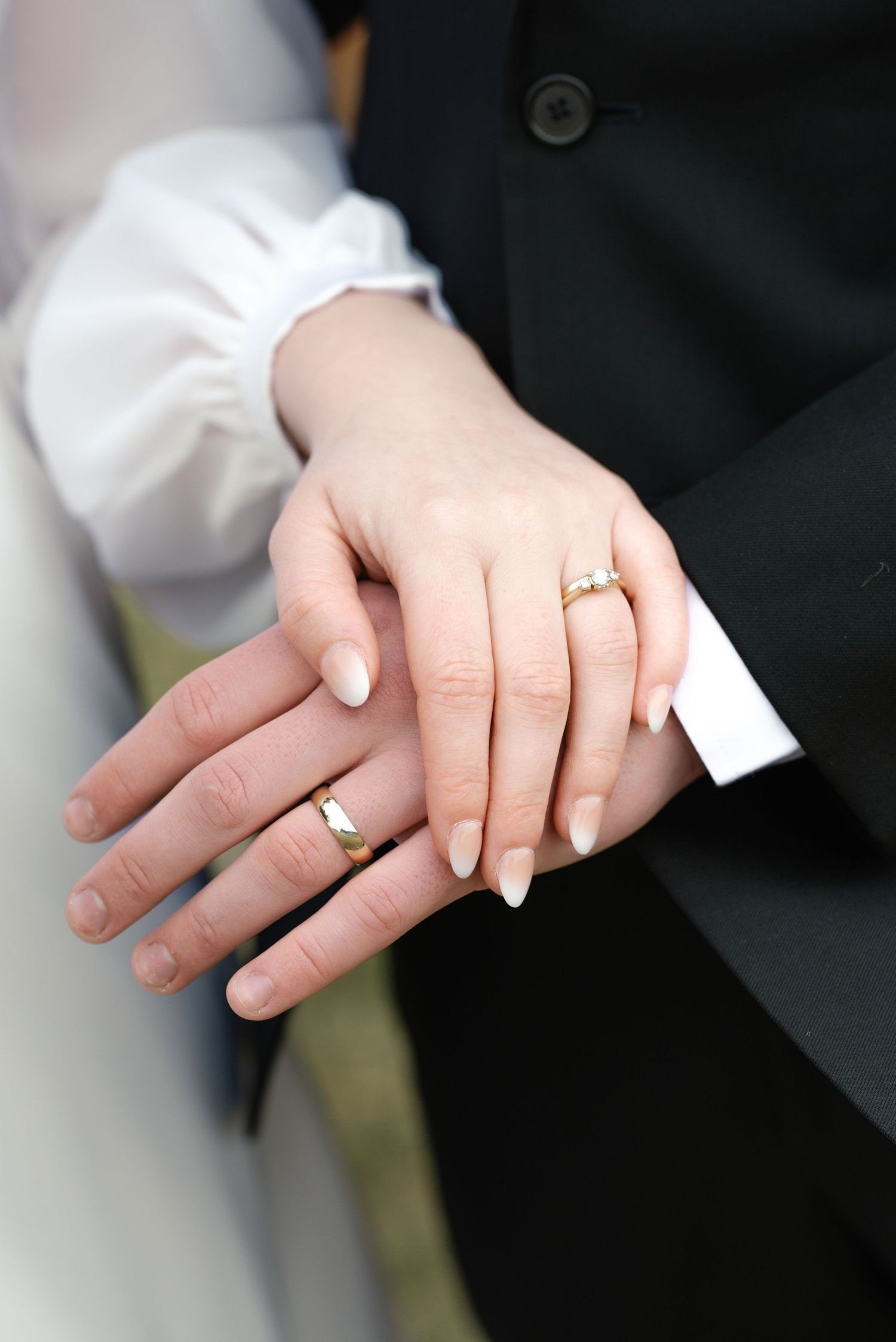 Newlyweds display gold wedding rings on their hands against groom's black suit jacket.