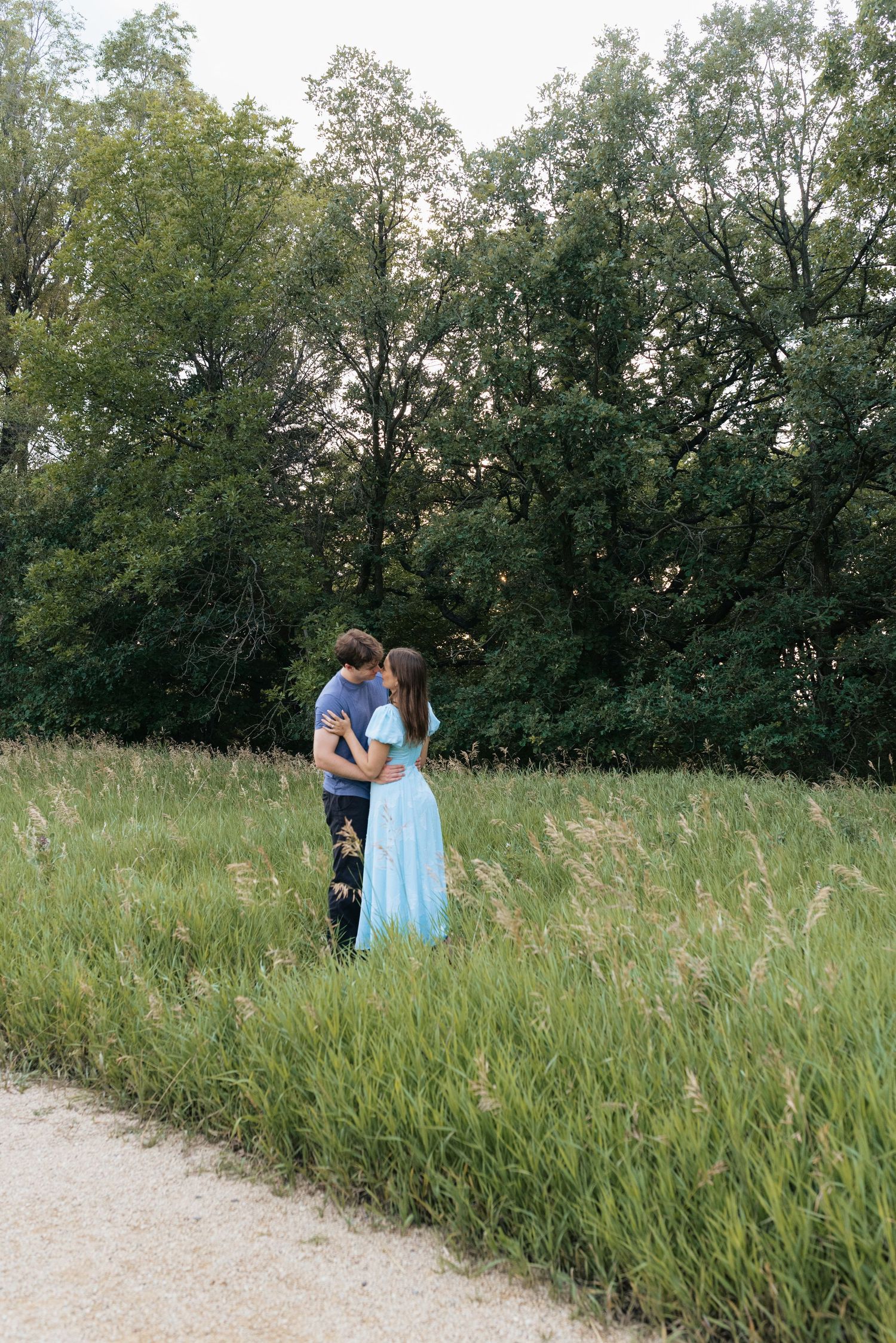 Couple embracing in a lush green meadow, woman in light blue dress, surrounded by tall grass and trees at dusk.