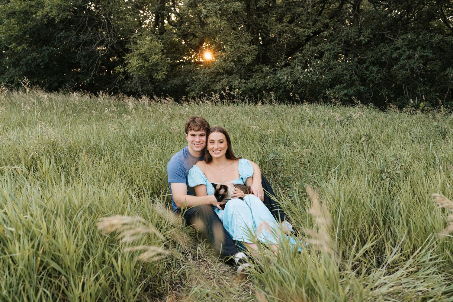 Couple sitting together in a lush green meadow at golden hour sunset, smiling warmly for an outdoor portrait.