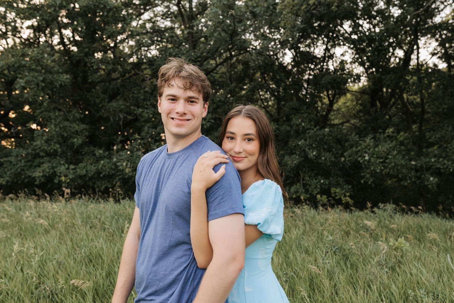 A young couple poses in a green meadow, the woman in a light blue dress resting her hand on the man's shoulder.