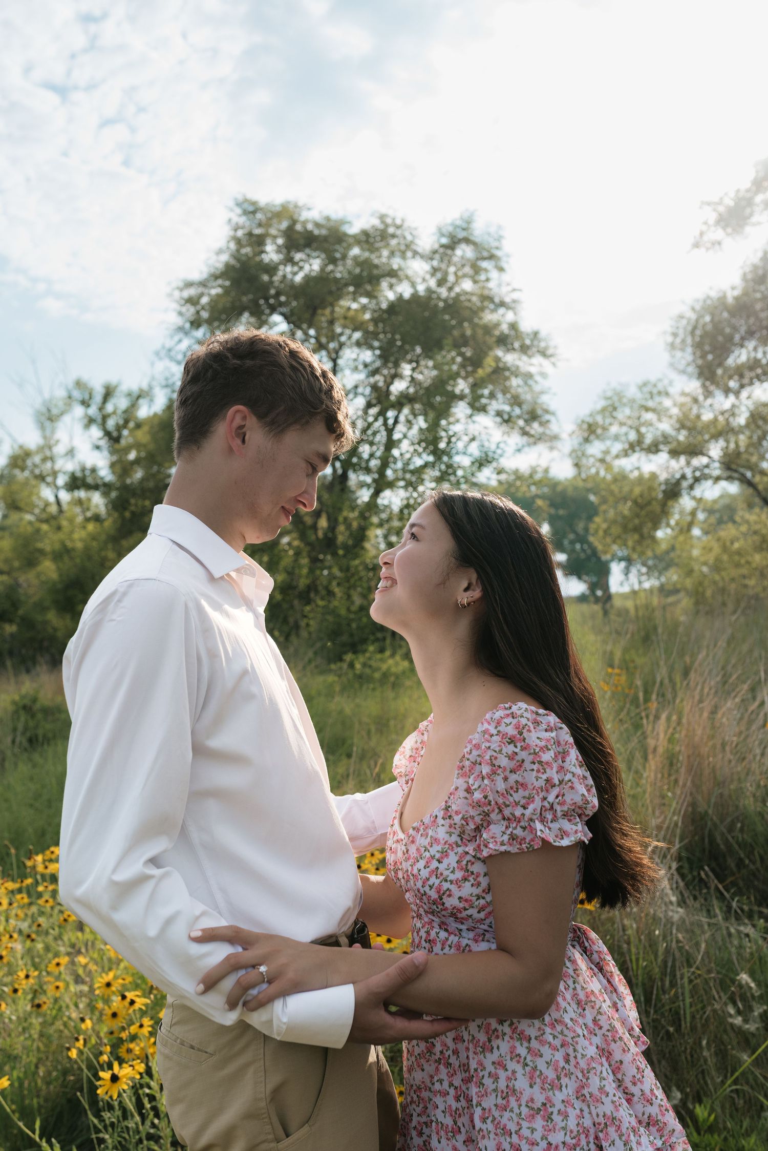 Couple gazing at each other lovingly in a sunny meadow with yellow wildflowers nearby.