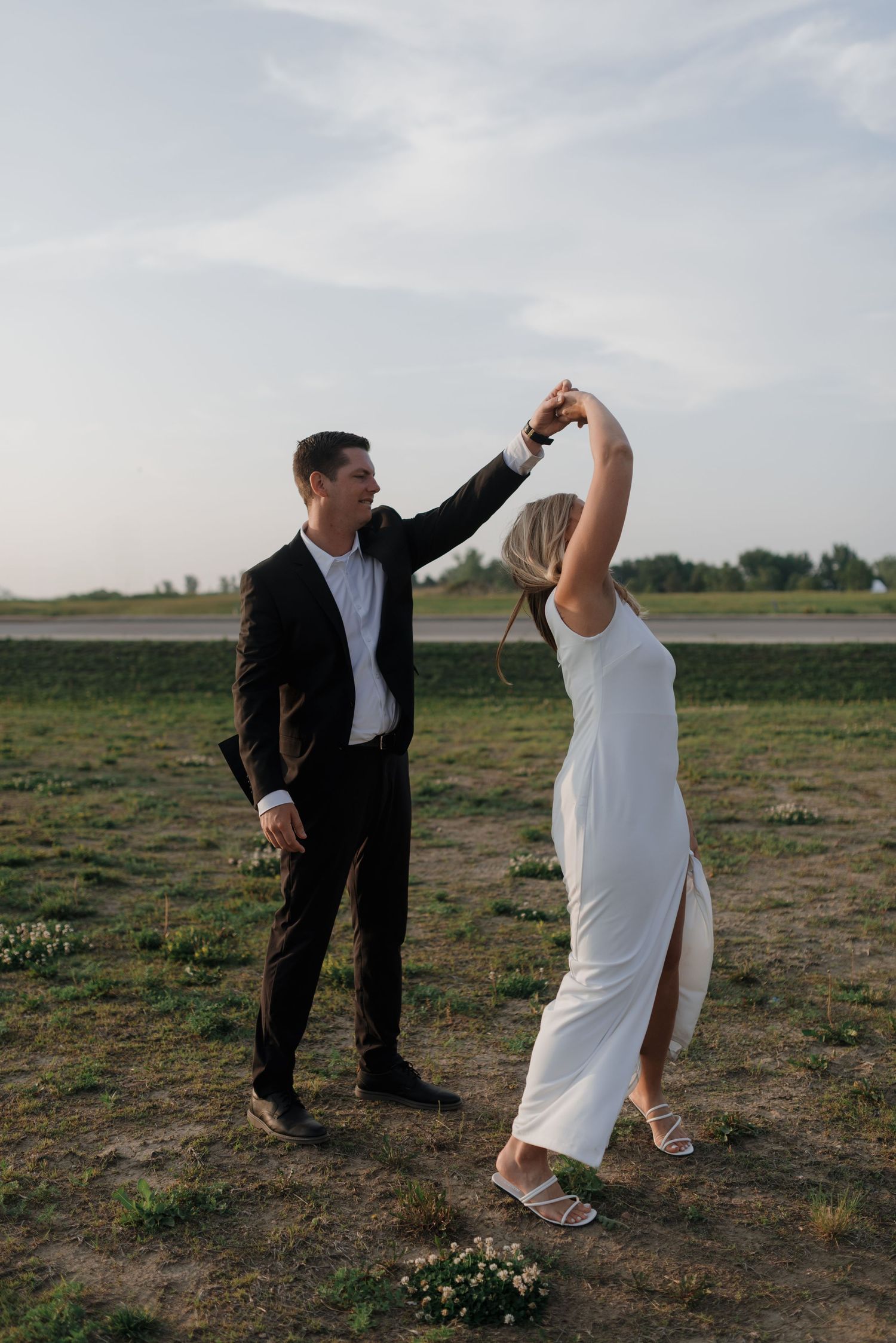 Groom twirls bride in elegant white gown during outdoor wedding dance in green field.
