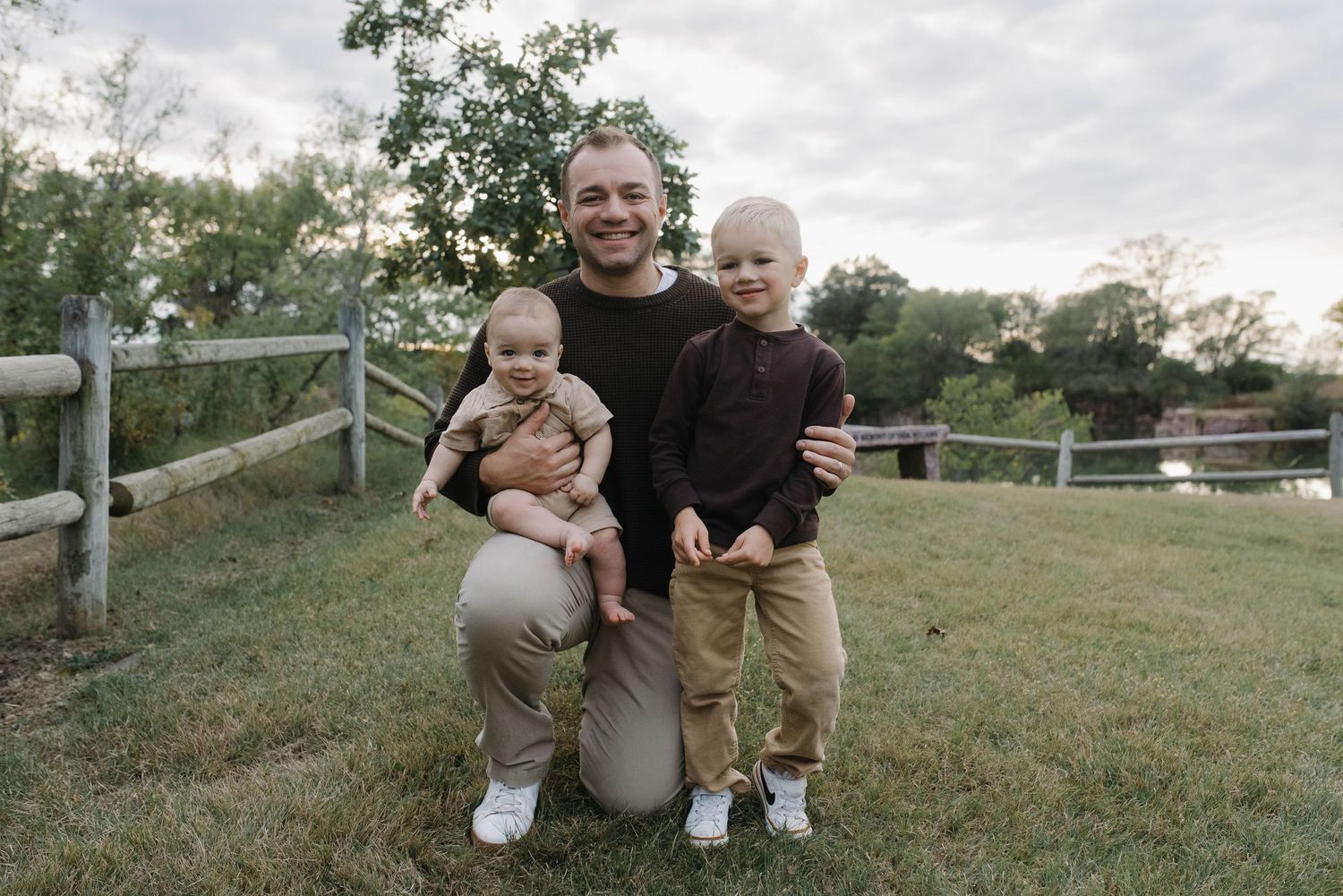 Father kneeling on grass holding baby while young toddler son stands beside him outdoors.