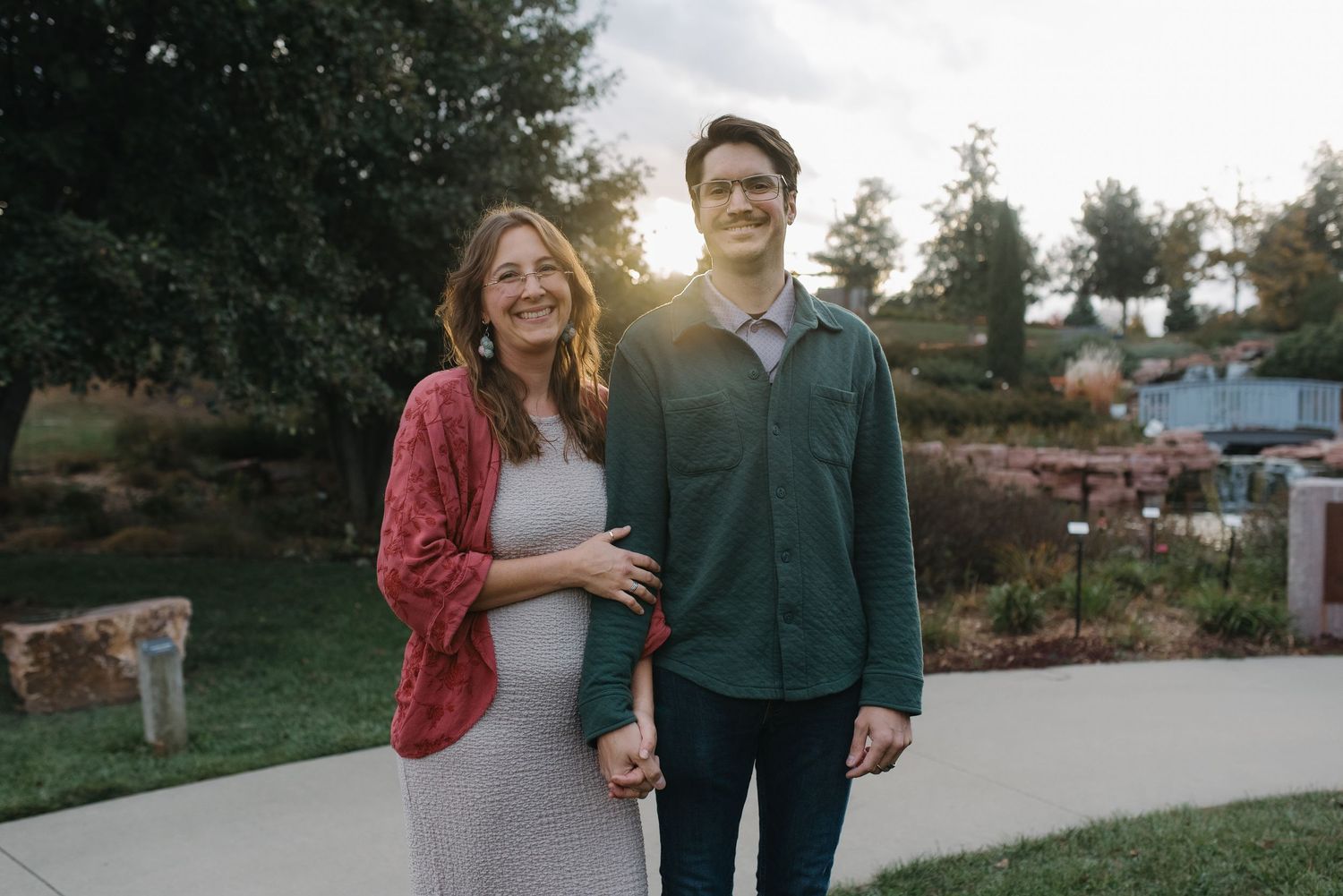 A smiling pregnant couple holds hands at sunset in a lush garden setting.