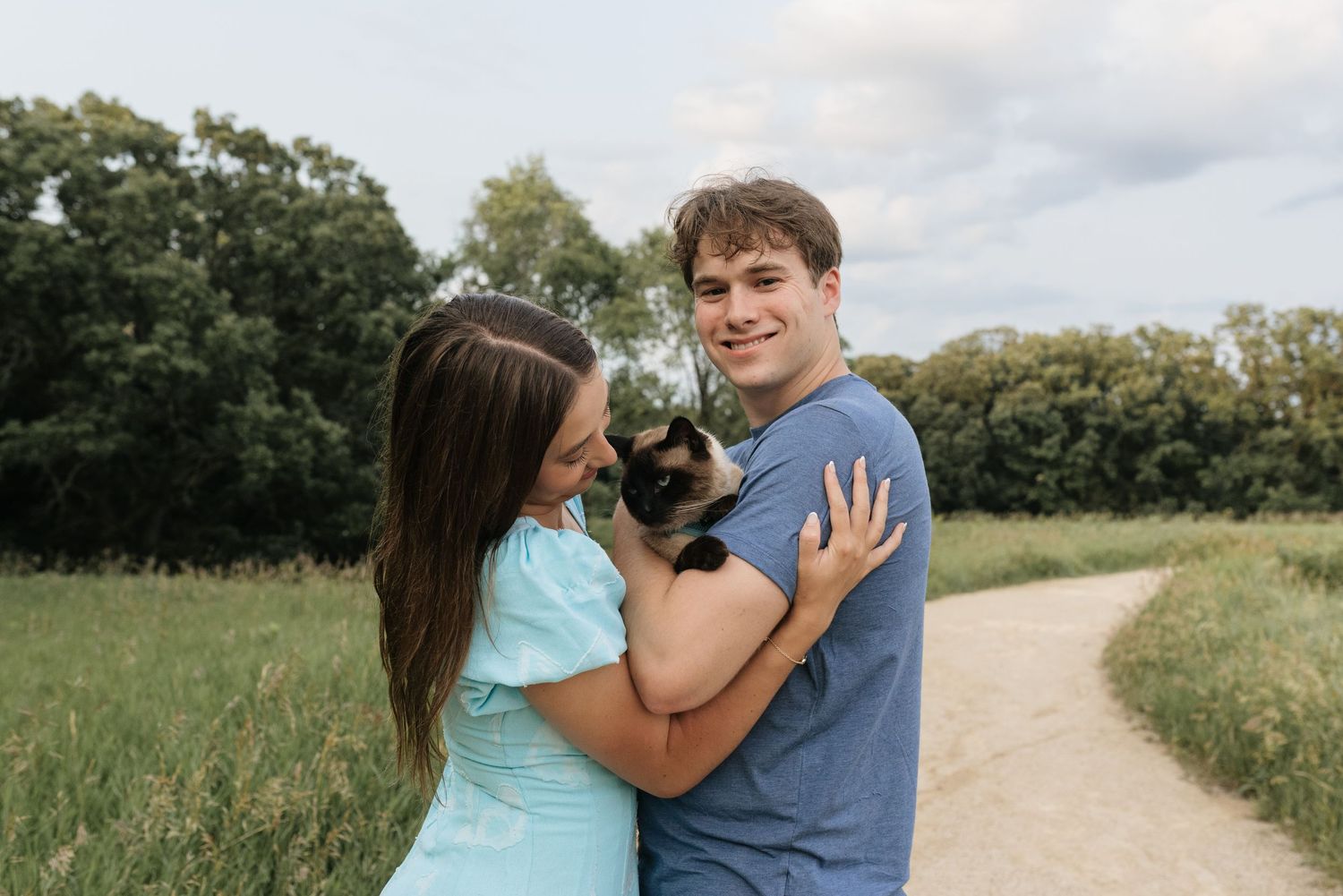 A couple smiles outdoors on a path, holding a Siamese cat, surrounded by green trees and grass.