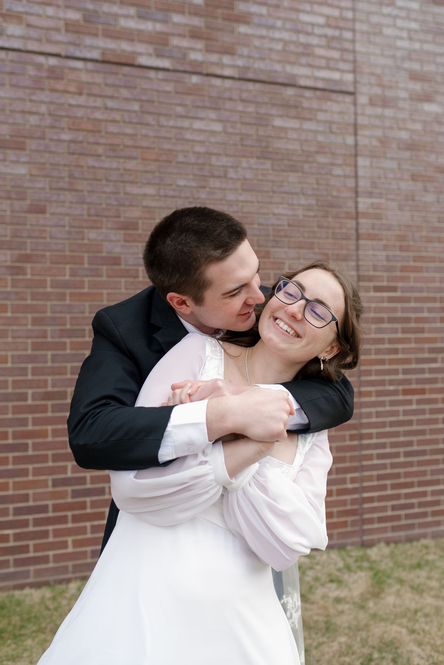 Smiling bride in white dress and glasses embraced by groom in black suit outdoors near brick wall.