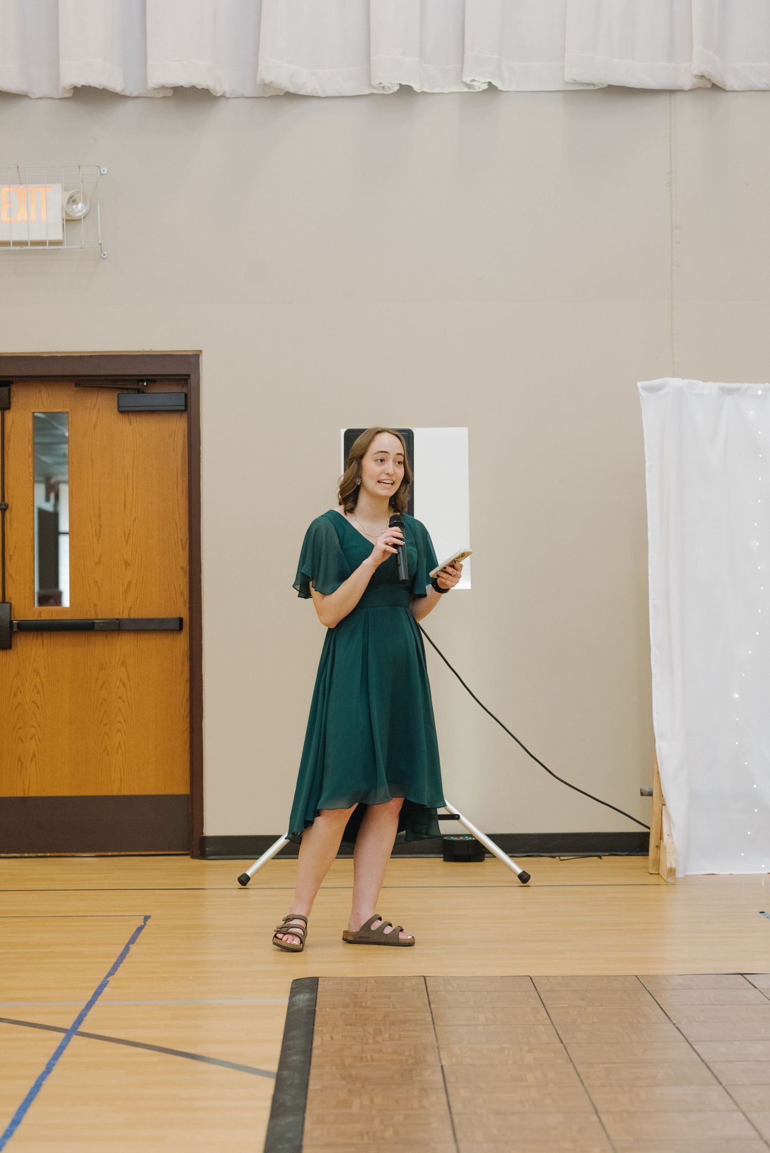 Woman in green midi dress speaks into microphone while holding phone in gymnasium setting.