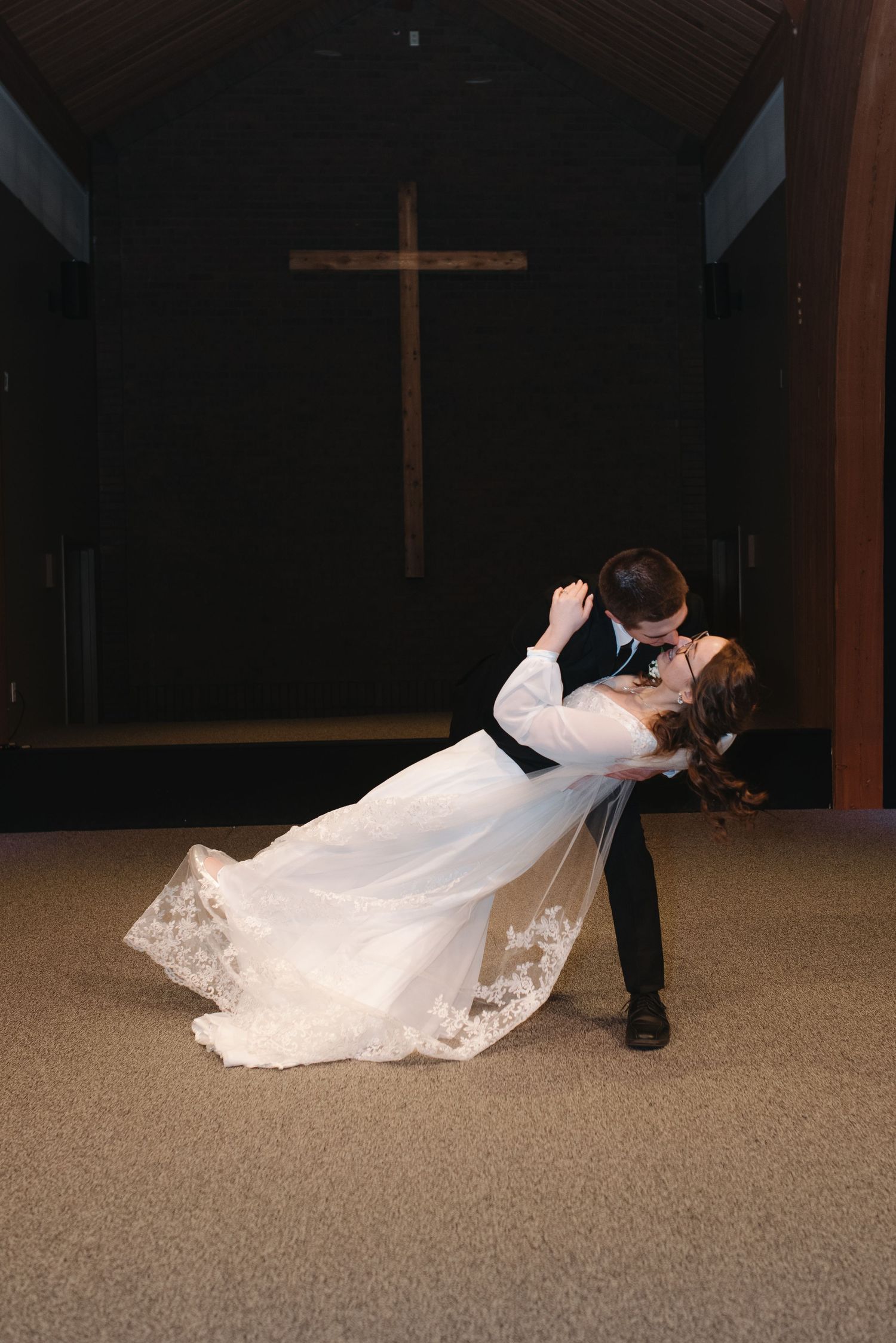 Groom dips bride dramatically in church aisle with wooden cross visible in background.