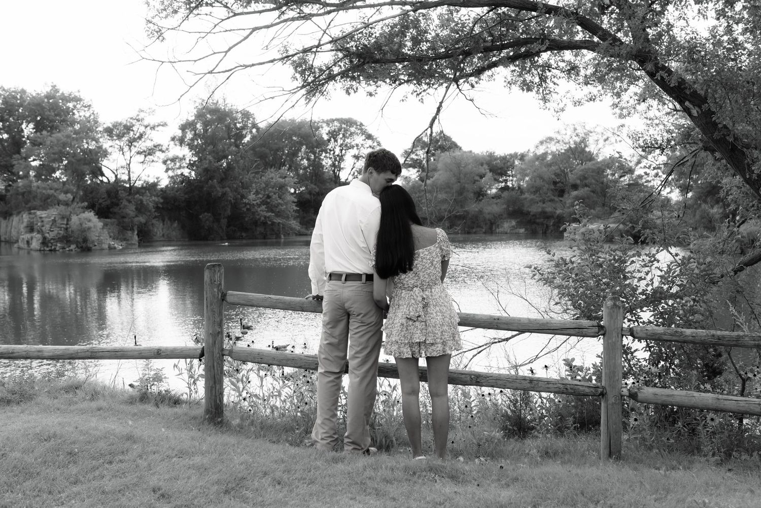 Black and white photo of a couple standing by a wooden fence overlooking a peaceful lake surrounded by trees.