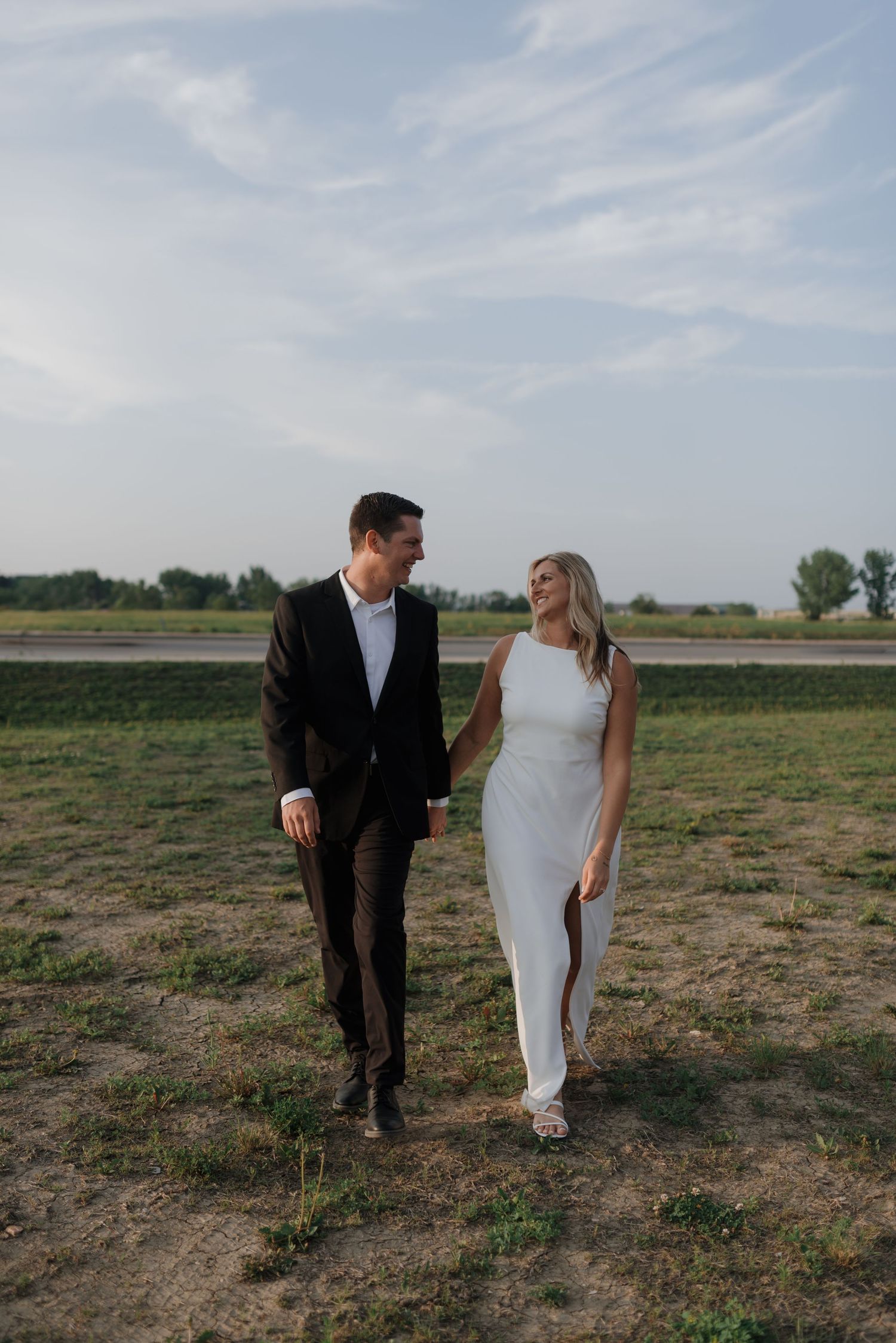 Elegant bride and groom holding hands and smiling while strolling through a green field.