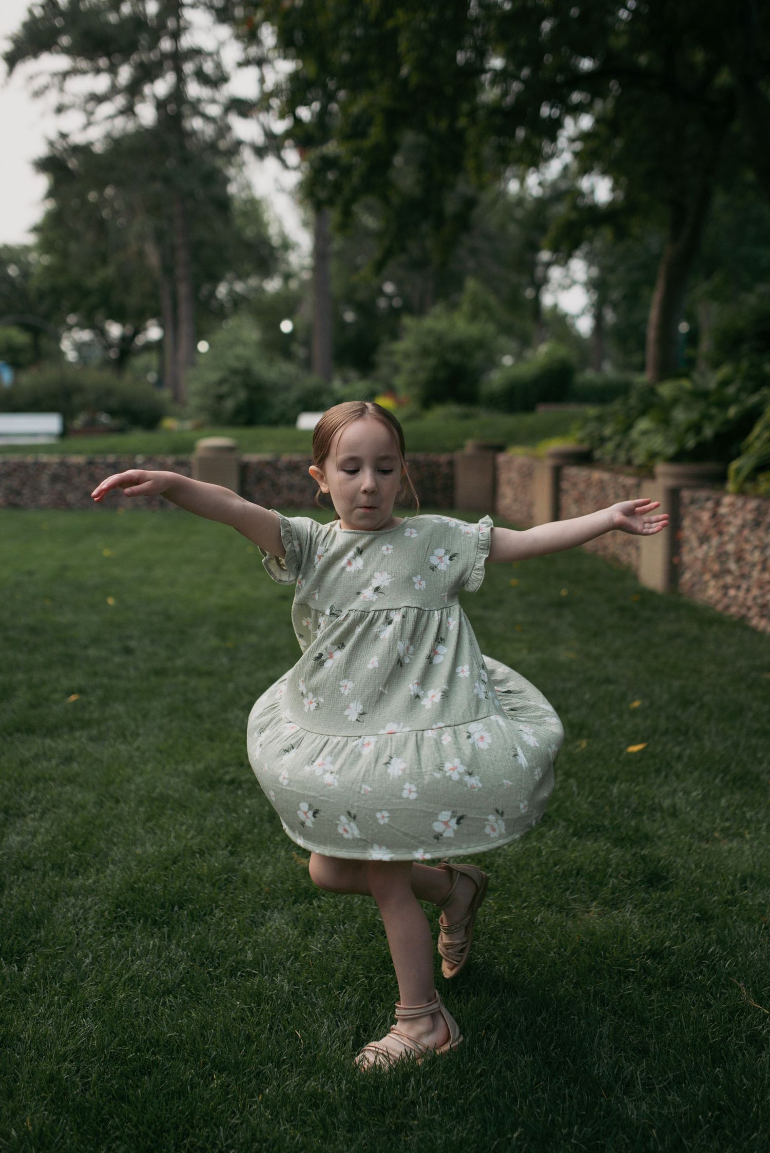 A young girl in a mint green floral dress pirouettes gracefully on one foot on green grass in a park.