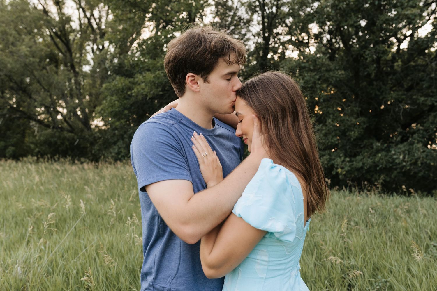 A couple shares a tender moment in a lush green field, the man gently holding the womans face as they lean in for a kiss.