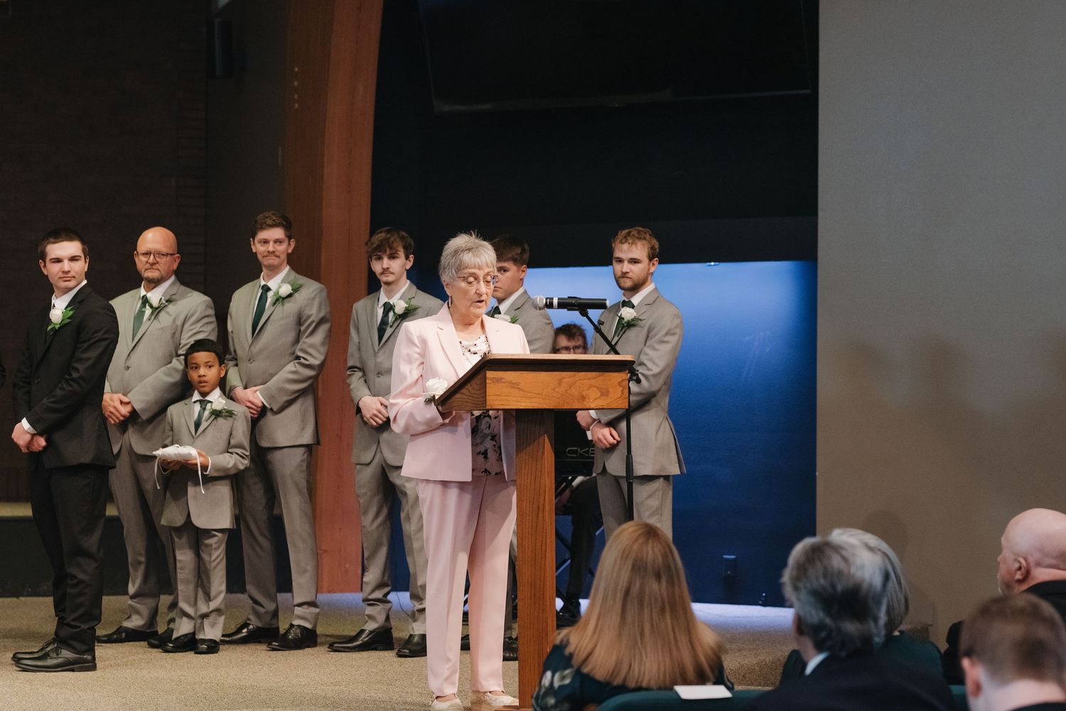 Elderly woman speaking at podium during formal ceremony with group of men in gray suits standing behind her.