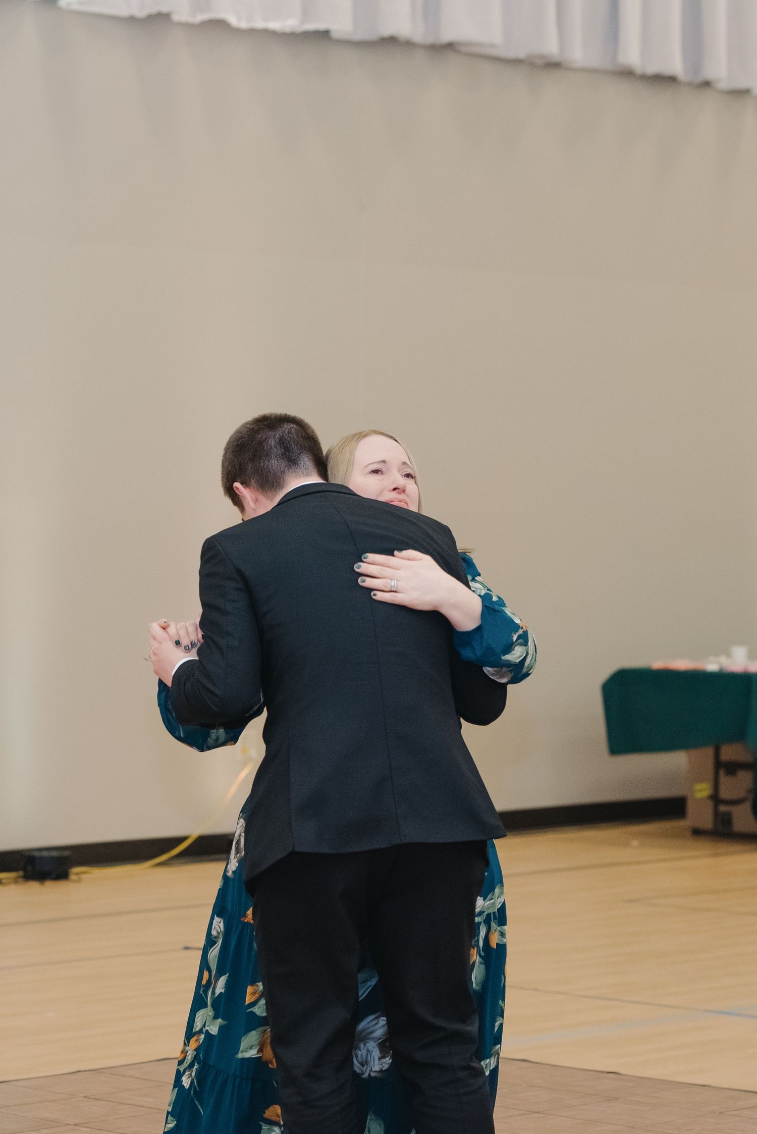 Bride in white dress sharing a warm hug with woman in teal floral dress on the dance floor.