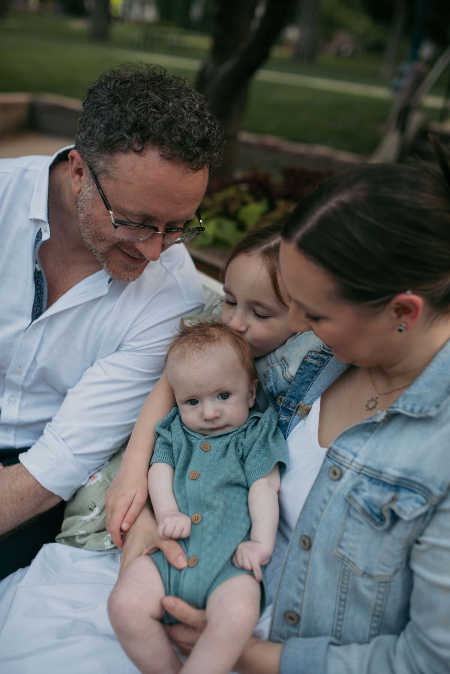 Family of four with baby in teal romper sharing a tender moment on a bench.