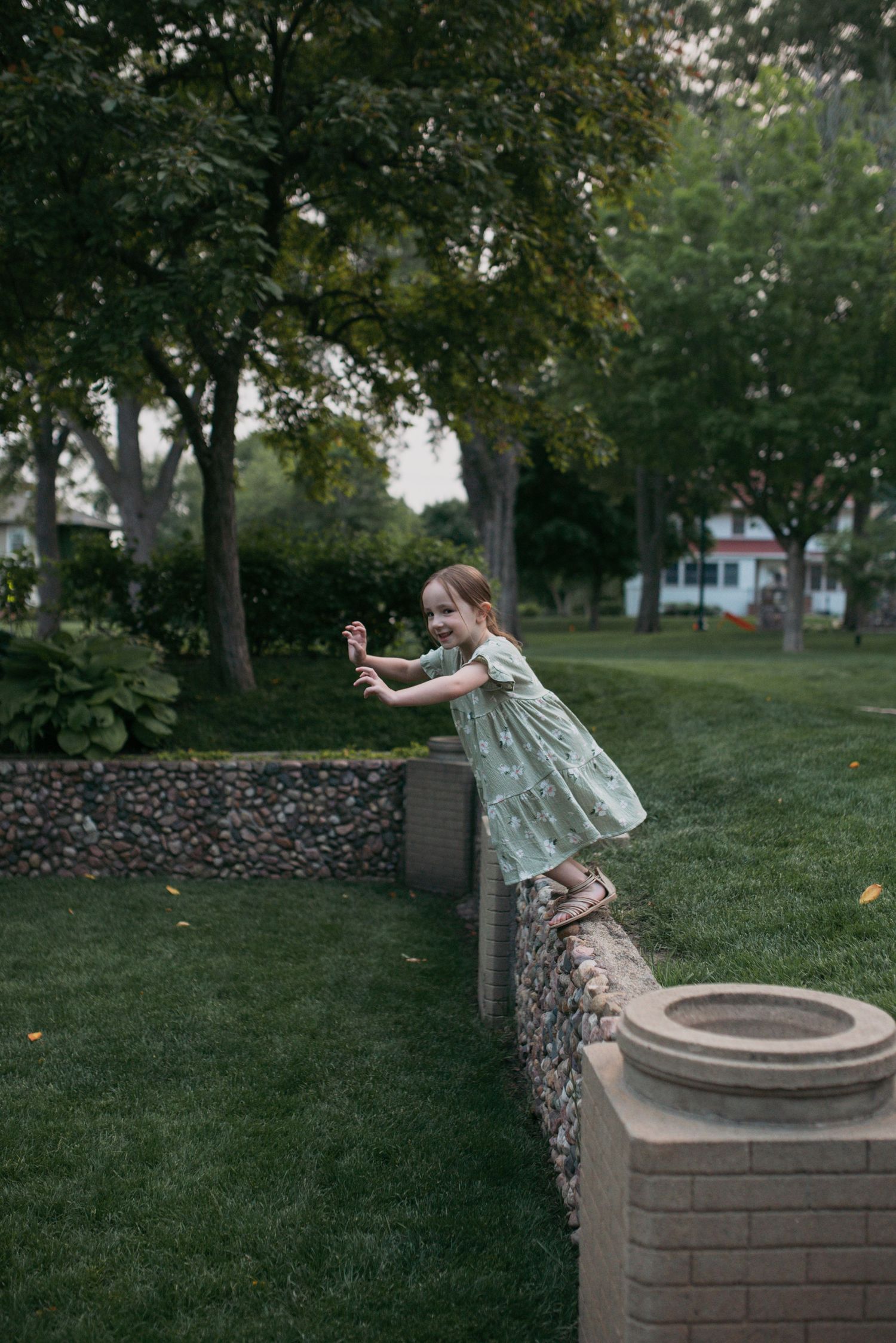 A playful young girl in a mint dress leaning over a stone garden wall in a green park.