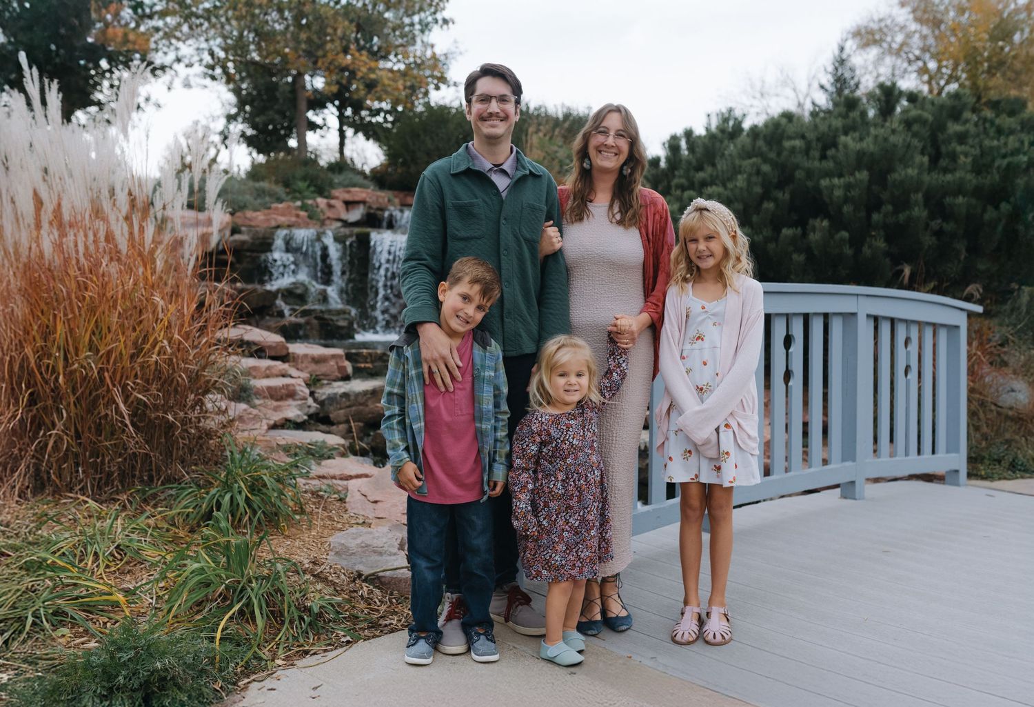Smiling family portrait with parents and three children on bridge near garden waterfall.