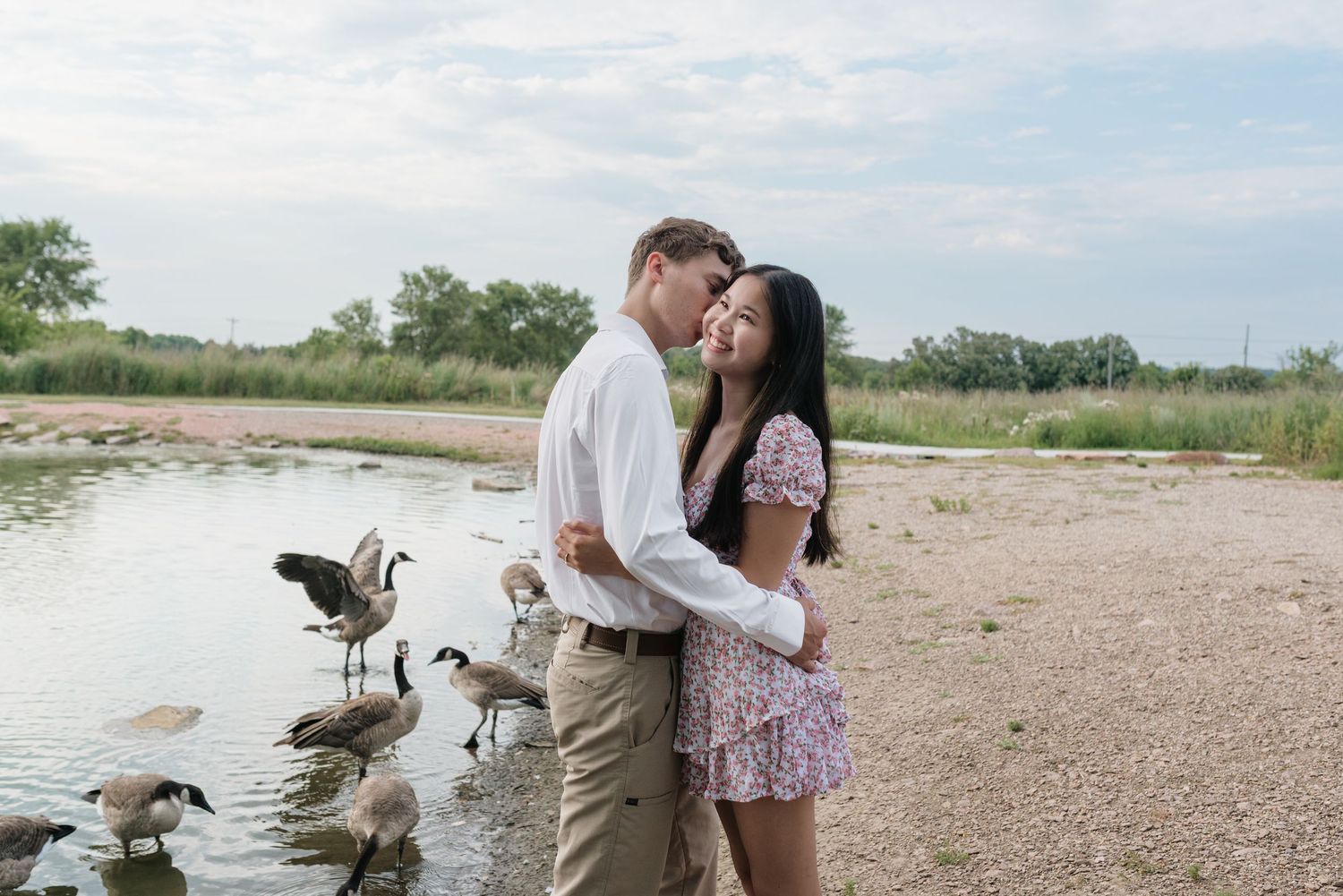 Couple embracing romantically by a lakeside with Canada geese nearby on a cloudy day.