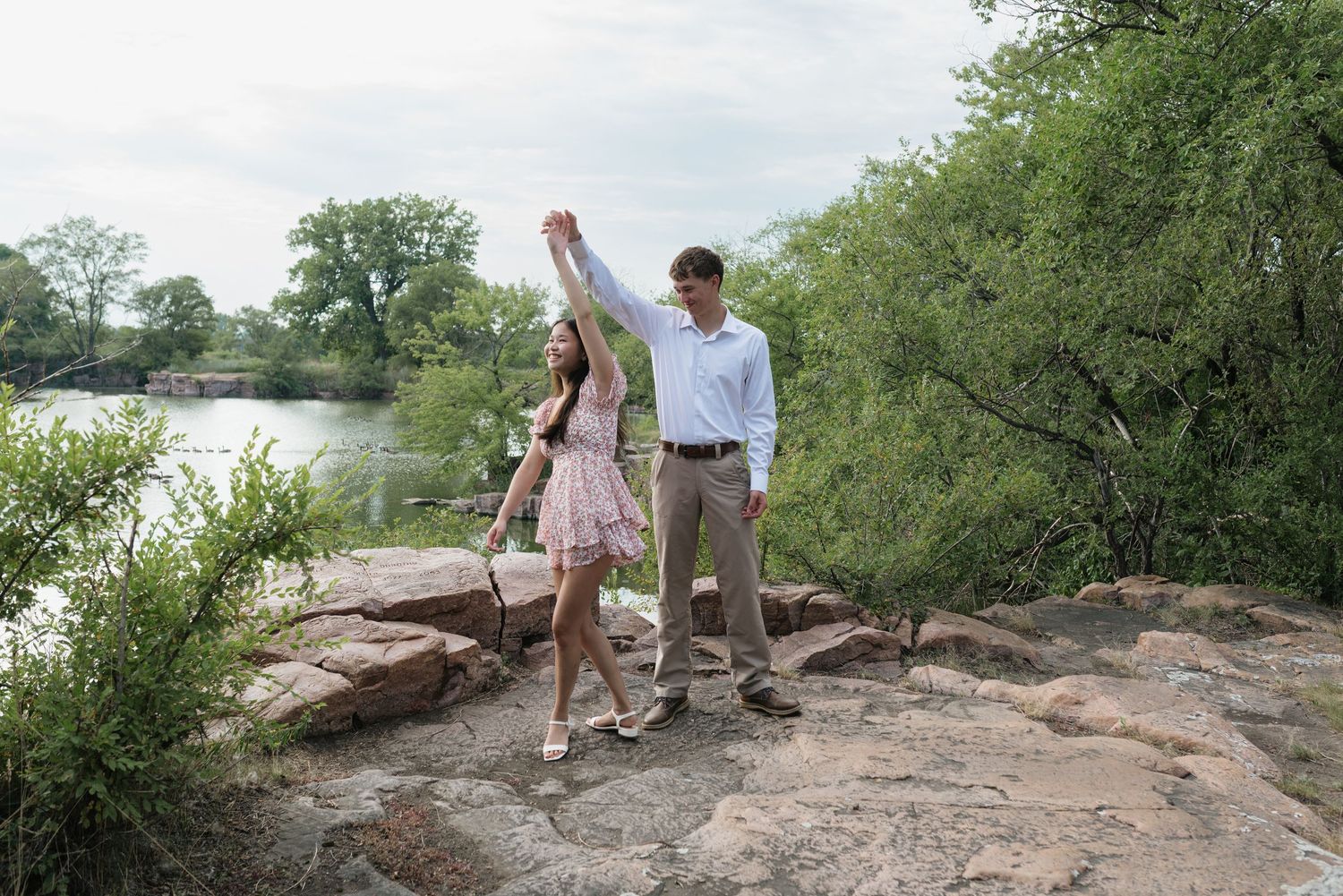 Couple dancing on rocky riverbank surrounded by lush green trees, man raising woman's hand in outdoor nature setting.