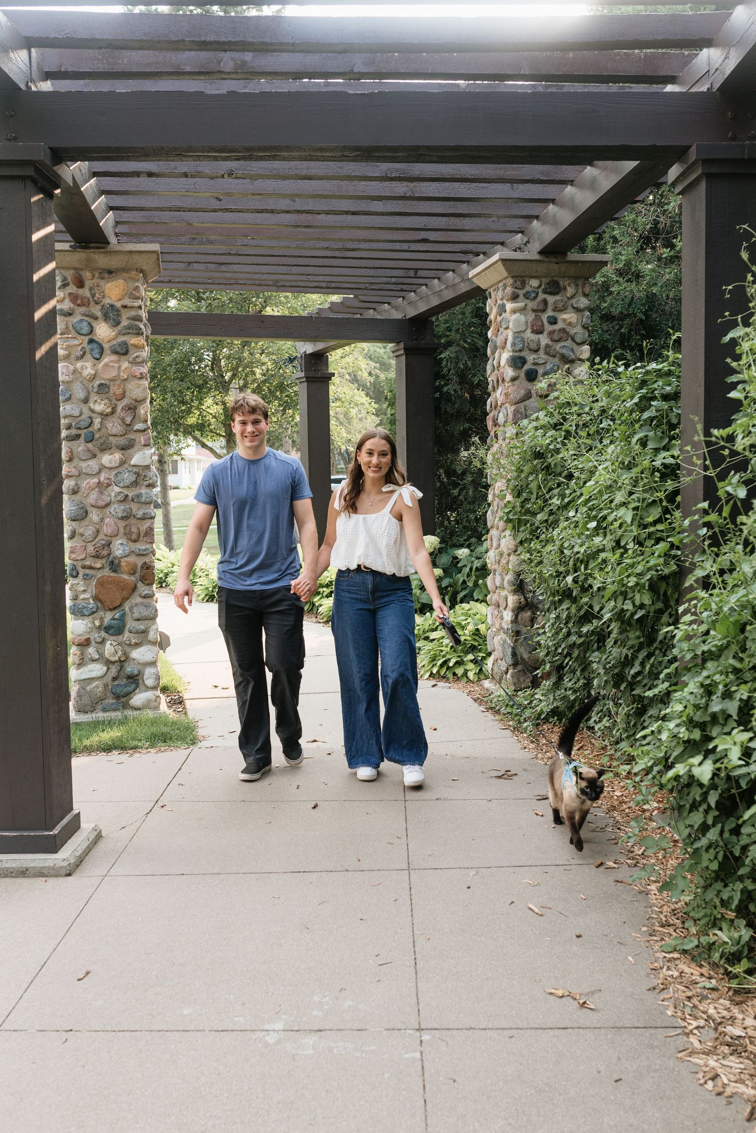 A couple walks hand-in-hand with a dog under a pergola surrounded by lush greenery on a sunny day.