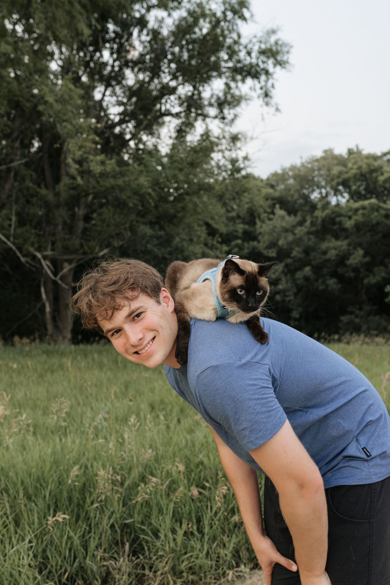 A smiling young man in a blue t-shirt leans forward with a Siamese cat perched on his back outdoors.