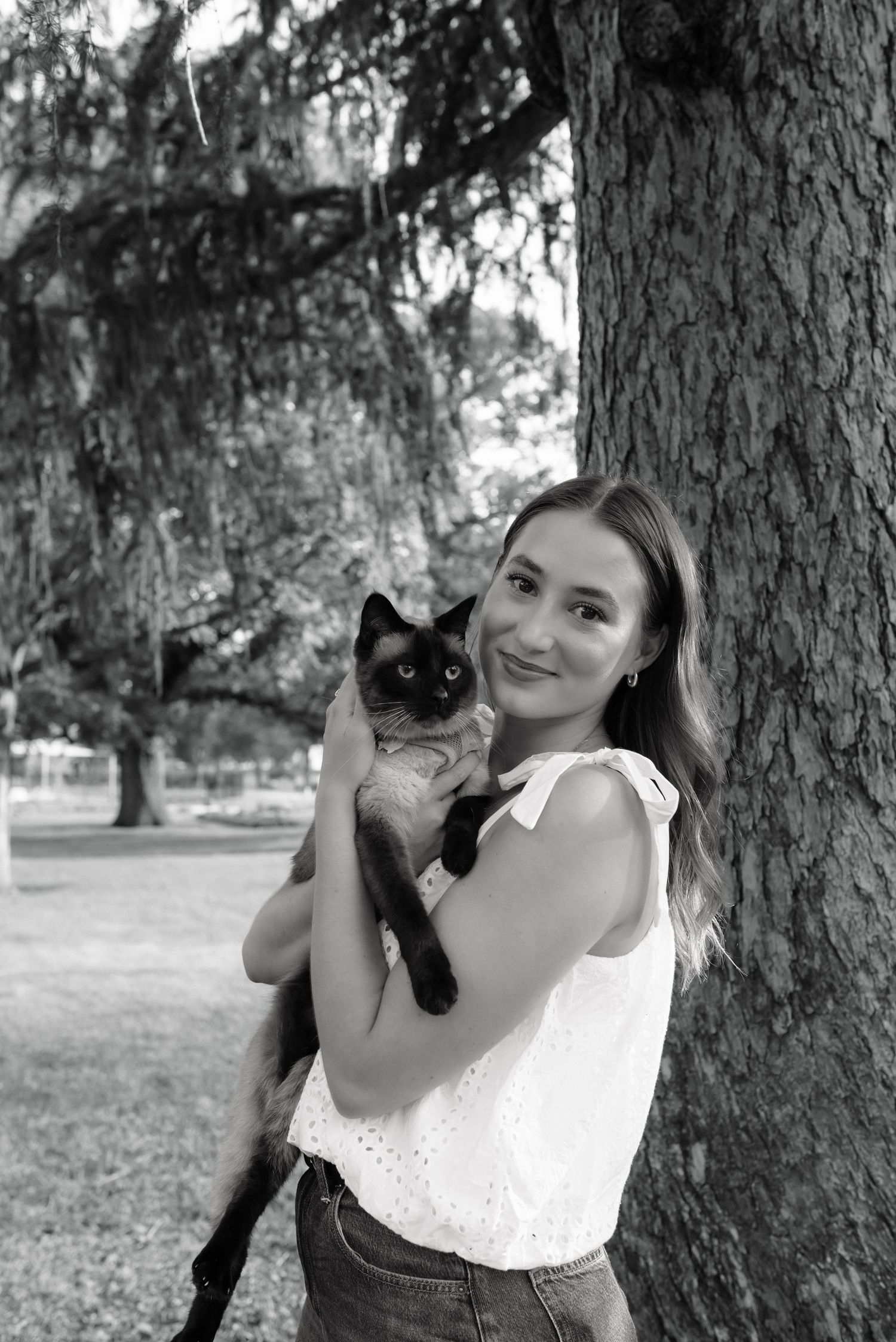 Young woman in white blouse smiles holding a Siamese cat outdoors near a large tree in black and white photo.