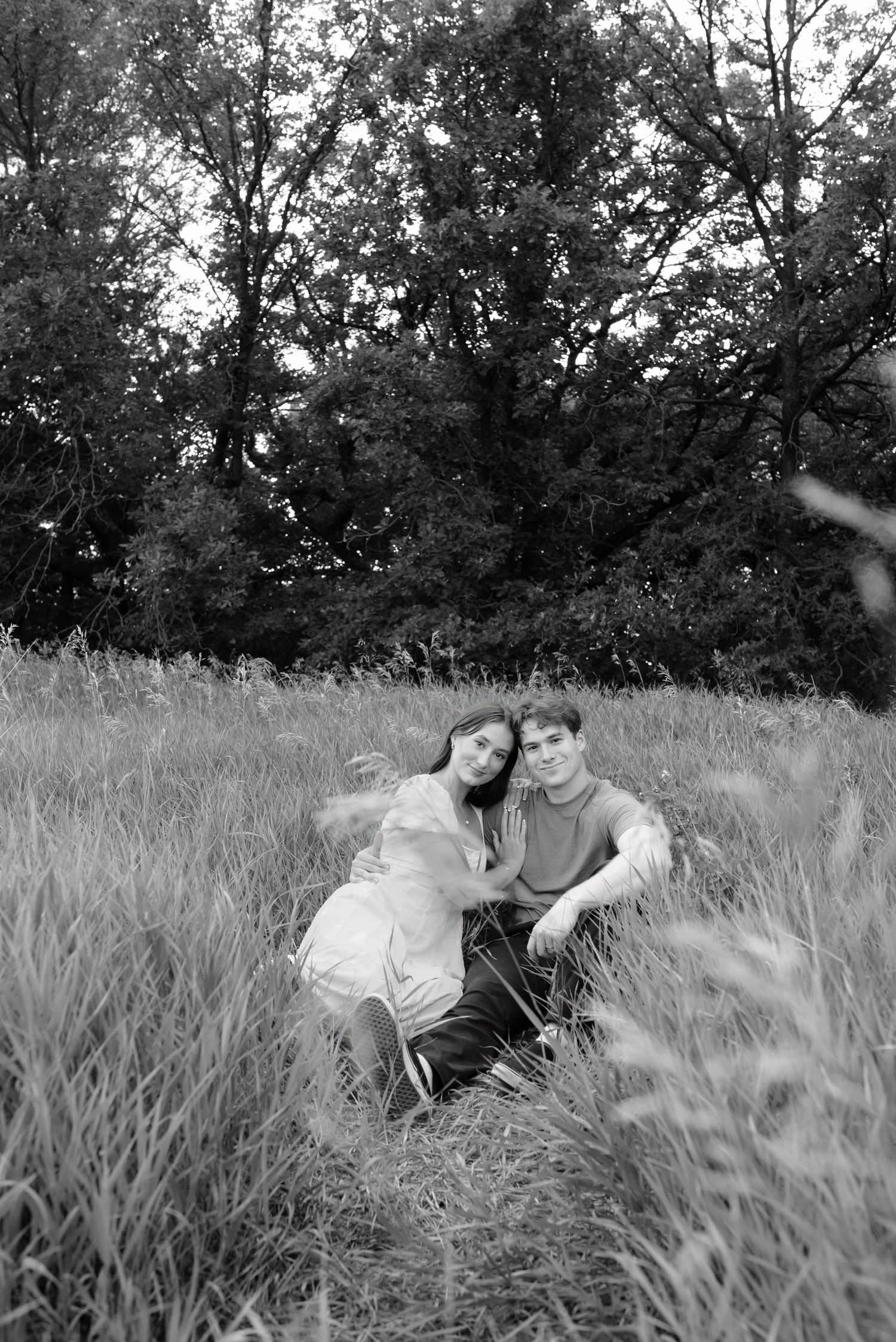 Black and white photo of a couple sitting together in a grassy field surrounded by trees.