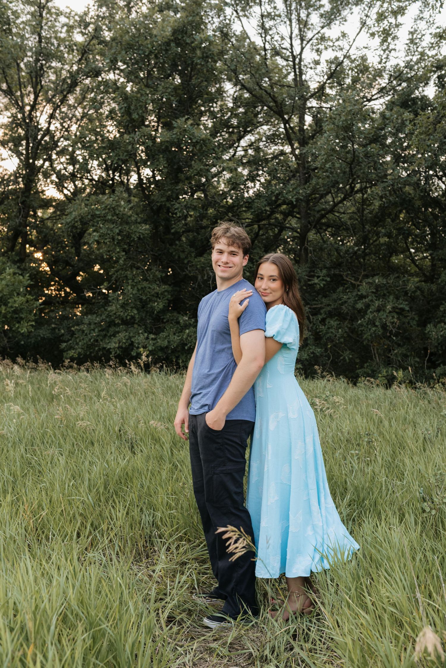 A young couple poses in a green meadow, she in a light blue dress, he in a blue shirt and black pants.