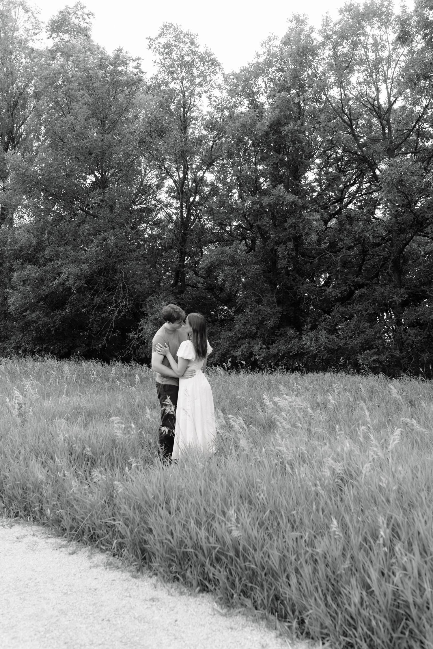 Black and white photo of a couple embracing in a lush green field with trees in the background.