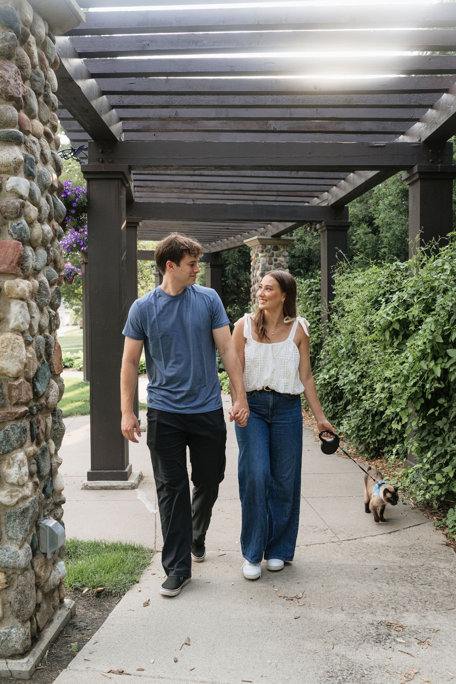 Couple holding hands and walking their dog under a pergola surrounded by lush greenery and stone pillars.