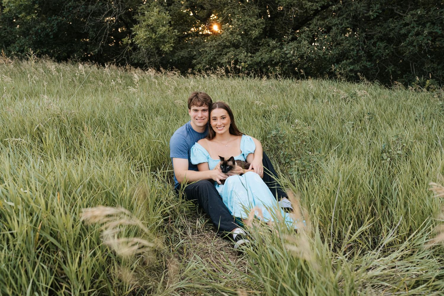 Couple sitting together in a lush green meadow at sunset, woman in blue dress holding a small dog.