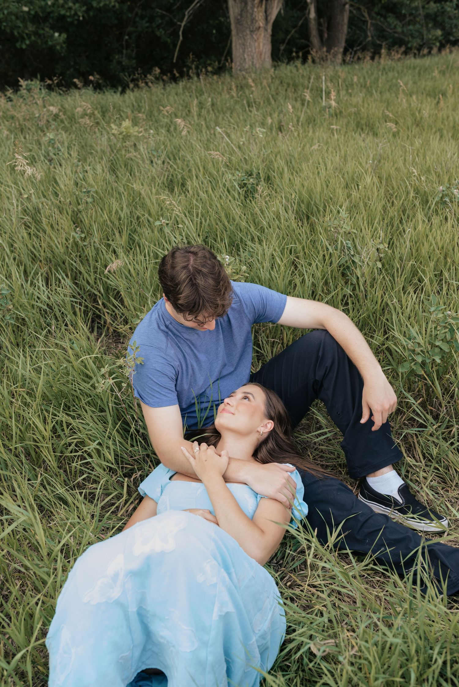 A couple relaxing in tall green grass, woman lying in man's lap smiling up at him in a serene outdoor setting.